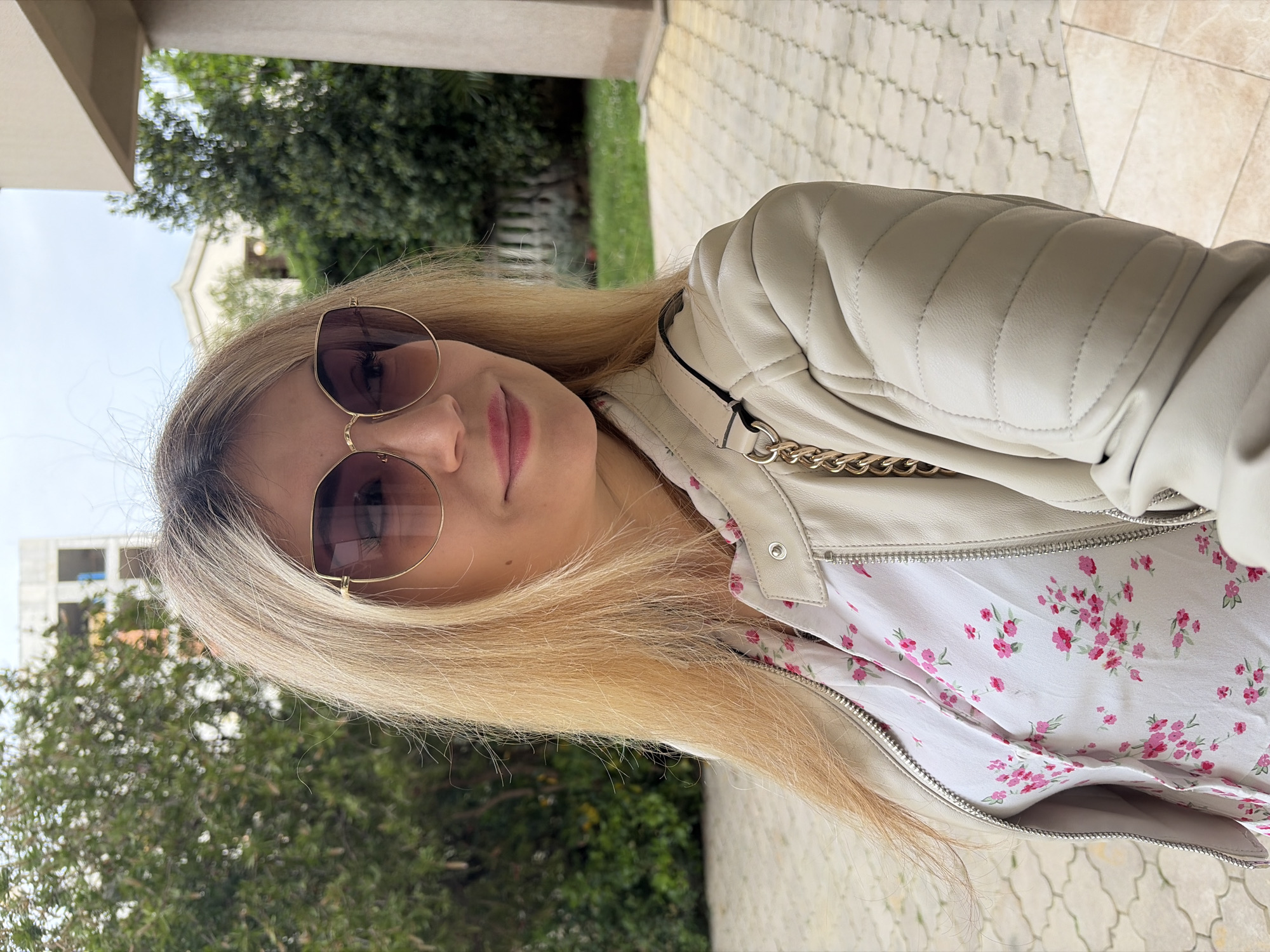 A blonde-haired woman wearing sunglasses is standing outdoors, likely in Budva, Montenegro. She is wearing a white leather jacket over a floral-patterned shirt. Behind her is a building under construction, trees, and stone paving. The woman is the main subject of the image, positioned in the foreground.