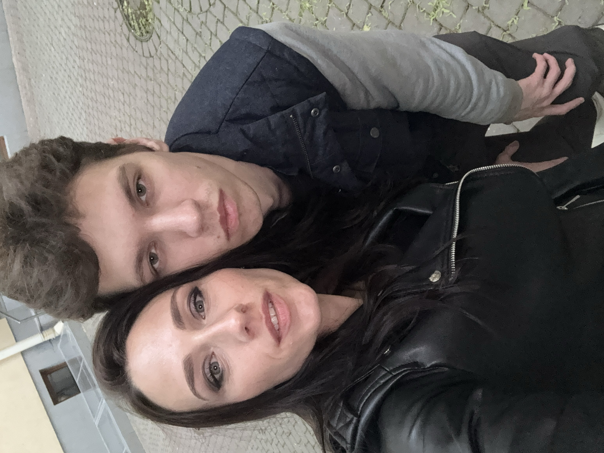 Two people, a man and a woman, are taking a selfie. The man is positioned slightly behind the woman. He has brown hair and is wearing a dark jacket with a gray hoodie underneath. The woman has dark hair and is wearing a black leather jacket. They are both looking at the camera. The background shows a paved surface, likely a sidewalk or square. A building is visible in the background. The location is likely Saint Petersburg, Russia, as the photo was taken there.