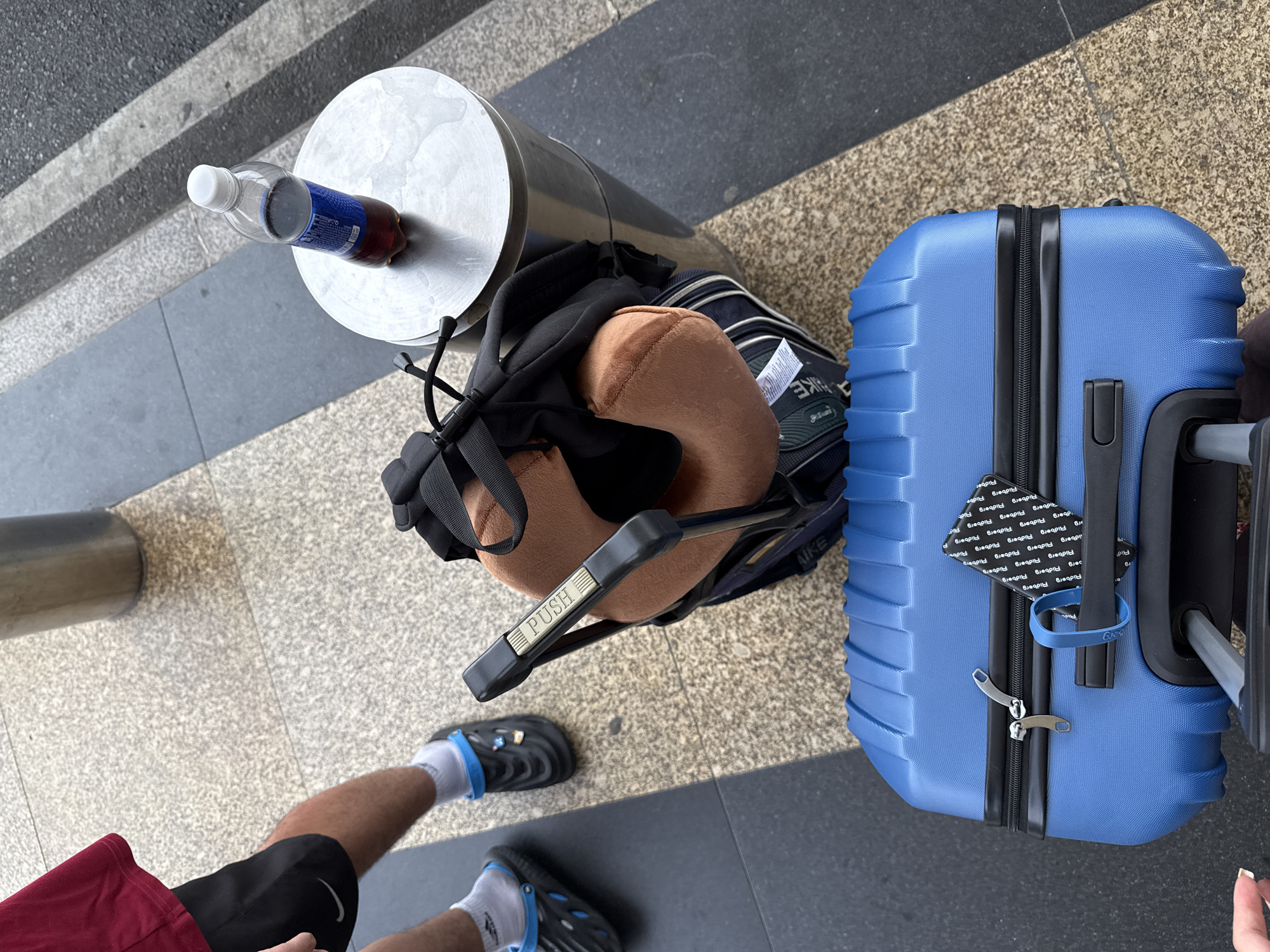 A high-angle view shows luggage and personal items on a patterned walkway, with a partial view of a person standing nearby. The person is wearing a red shirt, black shorts, white socks, and black sandals with blue accents. To the left, a silver cylindrical object, potentially a bollard, holds a plastic bottle of soda. A black backpack, topped with a tan neck pillow, rests on the walkway next to the bollard. The "PUSH" label is visible on the backpack's handle. To the right, a blue suitcase with a black handle stands upright, adorned with tags. The location appears to be an urban environment, suggested by the man-made walkway and the presence of street furniture. Based on the tags on the suitcase, the location may be Bac Cam Ranh Ward, Vietnam.