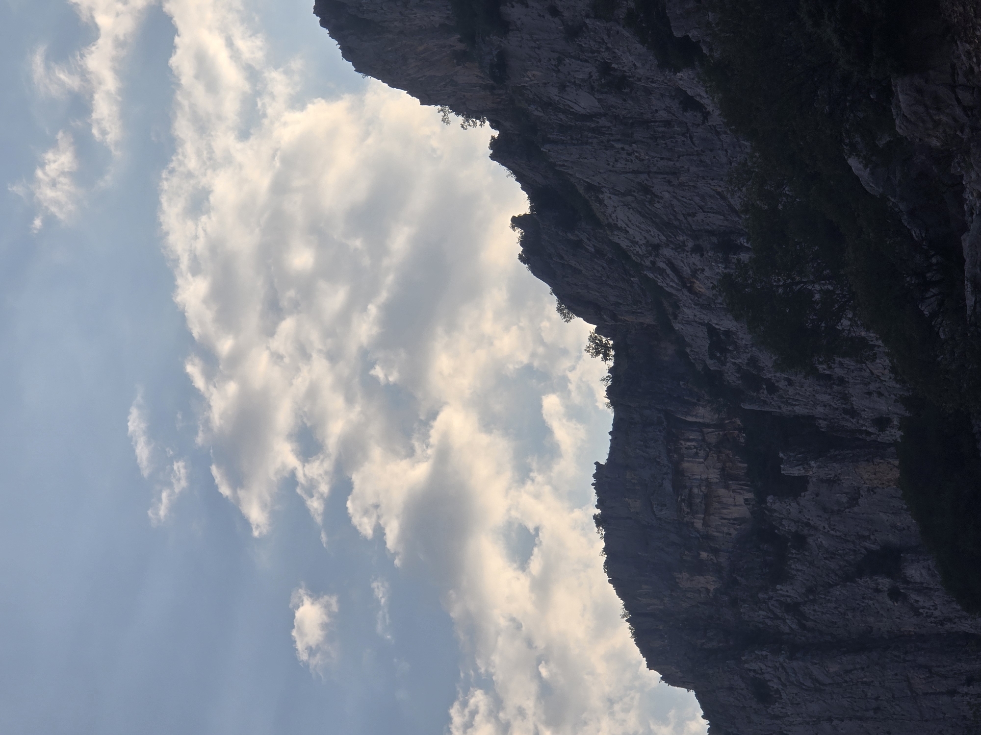 The image shows a dramatic view of a steep, rocky mountain range against a sky filled with dynamic clouds. The mountains are dark and textured, with visible rock formations and sparse vegetation clinging to their surfaces. The clouds are a mix of bright white and shaded gray, suggesting movement and light filtering through them, creating a sense of depth and atmosphere. The overall impression is one of natural grandeur and rugged beauty. The specific location in Podgorica, Montenegro is not identifiable from the image alone; it depicts a mountainous landscape common to the region. There are no people, animals, or man-made objects visible in the scene.