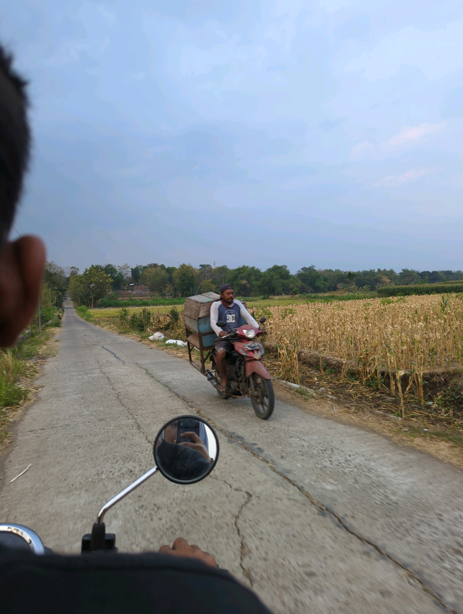 The image depicts a rural road scene in Senggrong, Indonesia. A man is riding a red motorcycle with a wooden cart attached, carrying several wooden boxes. He is wearing a blue t-shirt with white graphics and a dark cap. The motorcycle is on a cracked asphalt road that curves to the left and extends into the distance, lined by dry cornfields on the right and vegetation and trees on the left. The sky is overcast with muted blue and grey tones. In the foreground, the perspective suggests the photo is taken from another motorcycle, with a partial view of the rider's shoulder and a rearview mirror reflecting the rider's hand holding a mobile phone. The overall environment appears to be agricultural and serene.