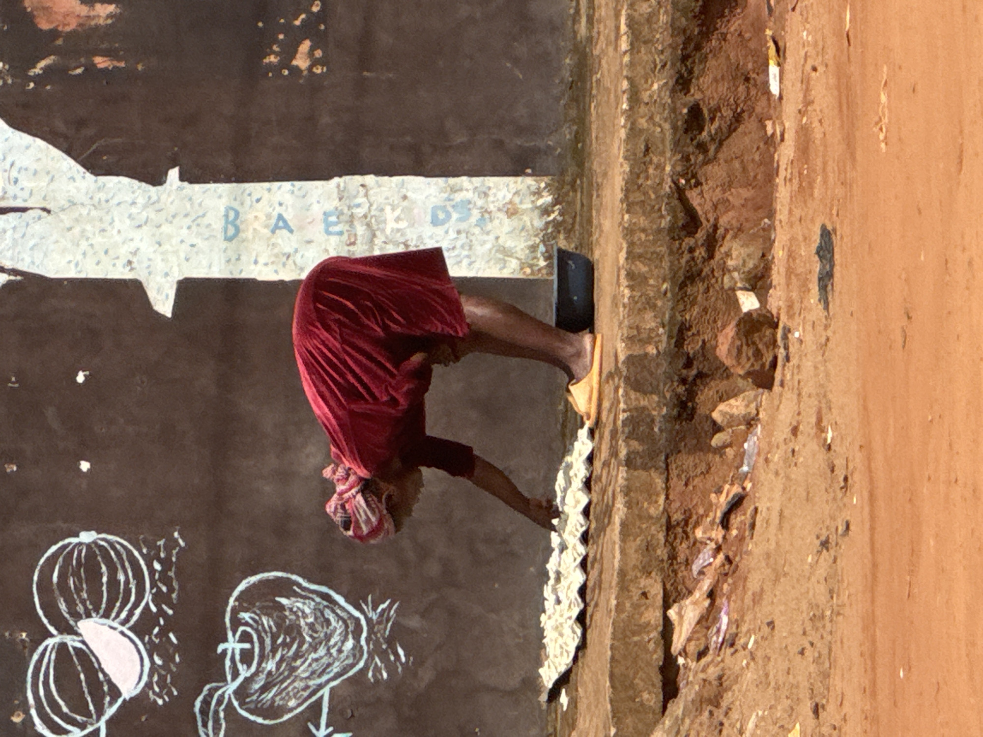 An elderly African woman in a red dress and headscarf is bent over, touching a textured white surface on a brown wall. She is wearing a yellow sandal on her visible foot and a black shoe on her other foot. The wall is decorated with white chalk drawings of fruits and the word "BAE" painted in blue letters above a white, irregular shape. The area adjacent to the wall appears to be a dirt path or road, with scattered debris and rocks. The setting is urban, likely in Onitsha, Nigeria, given the style of dress and architecture.