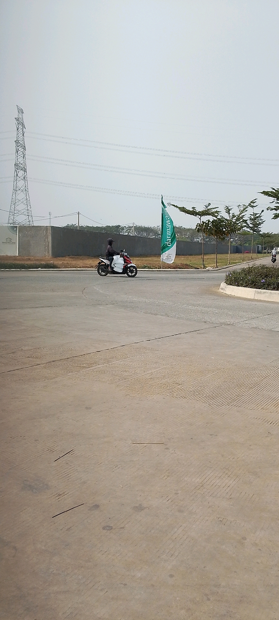 The image depicts a street scene in Satriamekar, Indonesia. In the mid-ground, a person wearing a dark hoodie is riding a white and red motorcycle. To the left of the motorcycle is a grey concrete wall with a sign that reads "DIWICITRA LAND". Further to the left, a tall, lattice-style power pylon stands against a hazy sky. To the right of the motorcycle, a green flag with "darmawa" printed vertically in white lettering is planted in the ground. Behind the wall and the flag, a row of young trees with sparse foliage is visible. The foreground is a paved road surface, with textured lines etched into the concrete. Another motorcycle is partially visible on the right side of the frame in the background, suggesting additional traffic. The overall environment appears to be an open, developing area with a mix of infrastructure and landscaping.