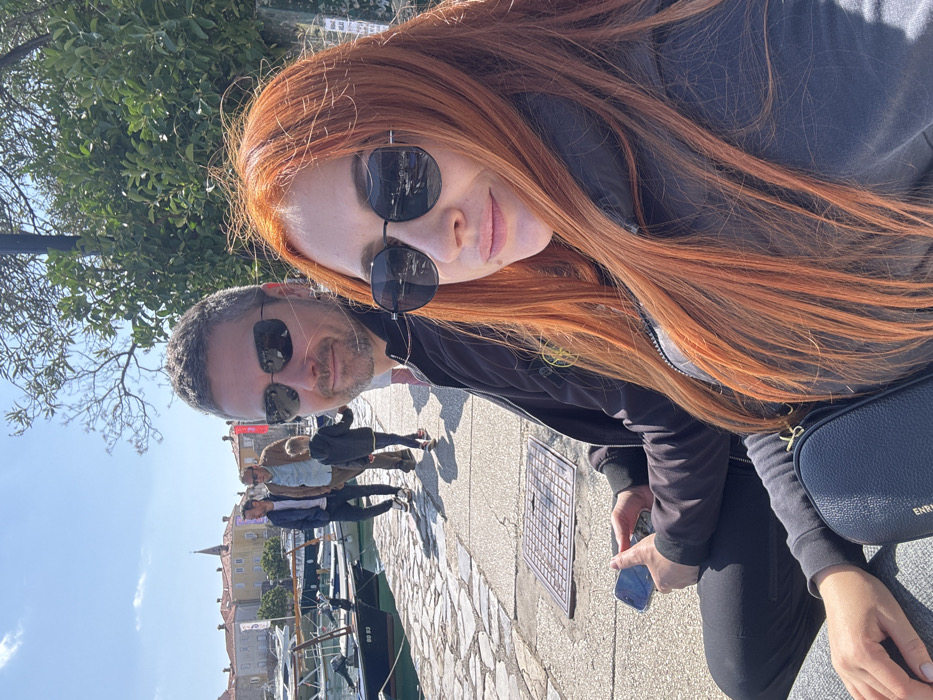 The image depicts a sunlit waterfront scene at the marina adjacent to Budva's Old Town in Montenegro. In the foreground, a woman with long, reddish-orange hair, wearing black sunglasses and a dark jacket, is positioned on the right, looking slightly to her right with a subtle smile. Behind her and to her left, a man with short grey hair, stubble, and dark sunglasses looks directly forward with a subtle smile; he holds a smartphone displaying a light-blue screen in his left hand. Both individuals are wearing dark jackets.
Behind them, on a light-colored cobblestone quay, three figures are walking away towards the left: two adult males, one in a blue jacket and another in a brown jacket, accompanying a young child in a dark jacket.
To the left of the quay, several small boats are moored in the water; one boat in the mid-ground prominently displays "BD 93" on its hull. The background features the historic architecture of Budva's Old Town, characterized by buildings with light-colored facades, red-tiled roofs, and a distinct church tower. Green foliage and bare tree branches are visible along the right side of the frame. A square metal grate is embedded in the cobblestone path in the mid-foreground. The sky is clear blue.