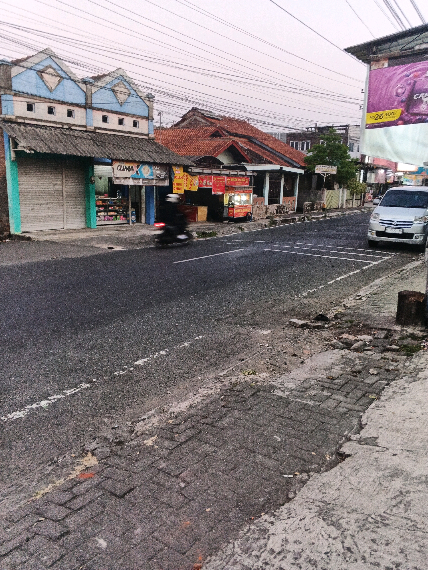 A street scene in Ungaran, Indonesia, shows buildings lining the right side of the road. The buildings include a store with a rolled-down metal shutter and a sign that says "CLIMAX KO". Next to it, another building has a red and yellow food stall with the words "BUNGKUS & GORENG" visible. Further down the road, a silver minivan is parked. The road itself is asphalt with white painted lines. The sidewalk on the right is paved with brick pavers and shows some signs of disrepair. A motorcycle is passing by, blurring its image. The sky is overcast.