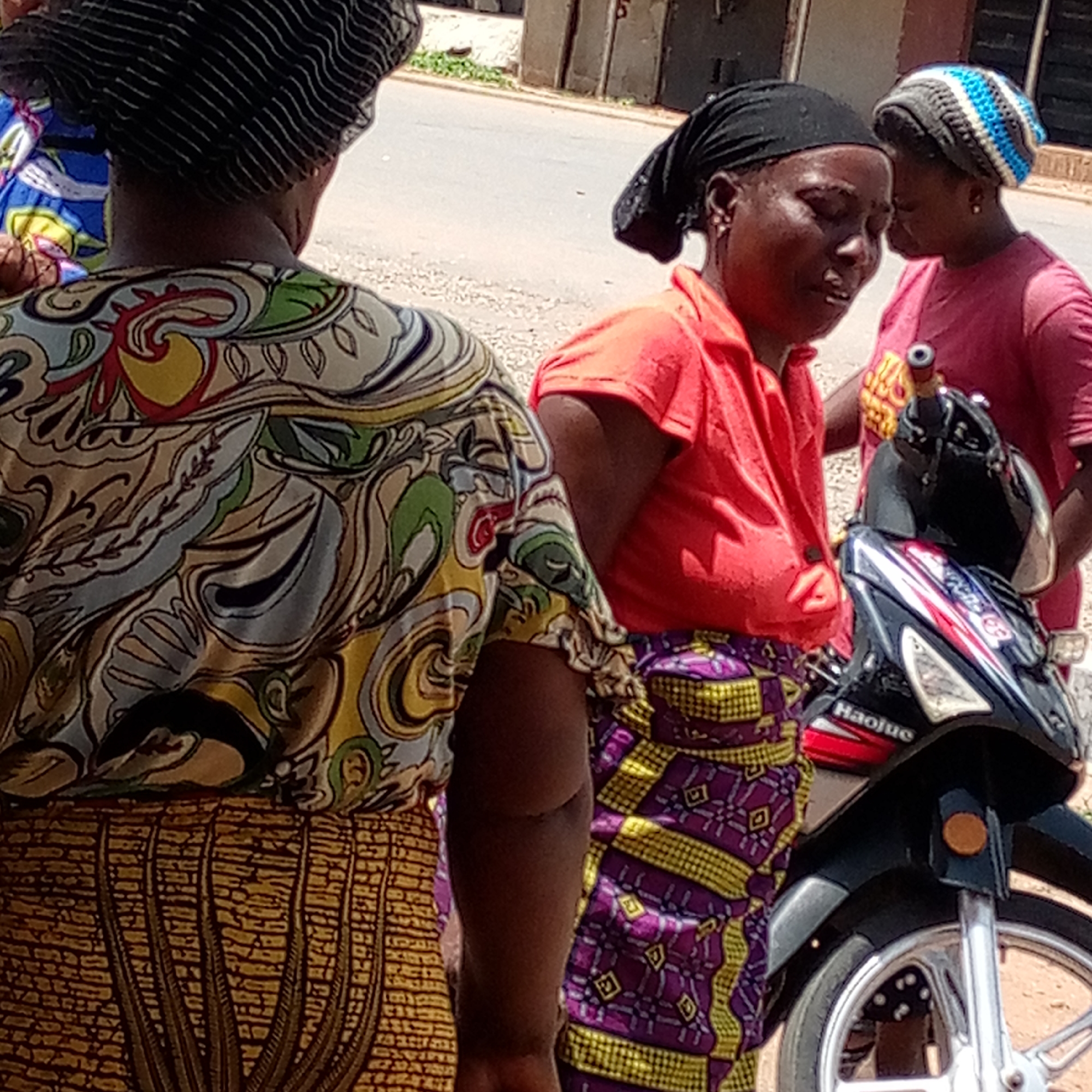 Three women stand next to a Haouje motorcycle in an outdoor setting in Mararaba, Nigeria. The woman closest to the camera has her back turned and wears a patterned yellow, brown, and green top and a brown skirt with a brick-like pattern. The woman in the center, standing to the right, wears a red top and a purple and yellow patterned skirt, along with a black headscarf. The third woman, partially visible behind the motorcycle and to the right of the woman in red, wears a red shirt and a blue and gray beanie. The motorcycle is primarily black with red, white, and blue accents. The background includes a road and buildings.