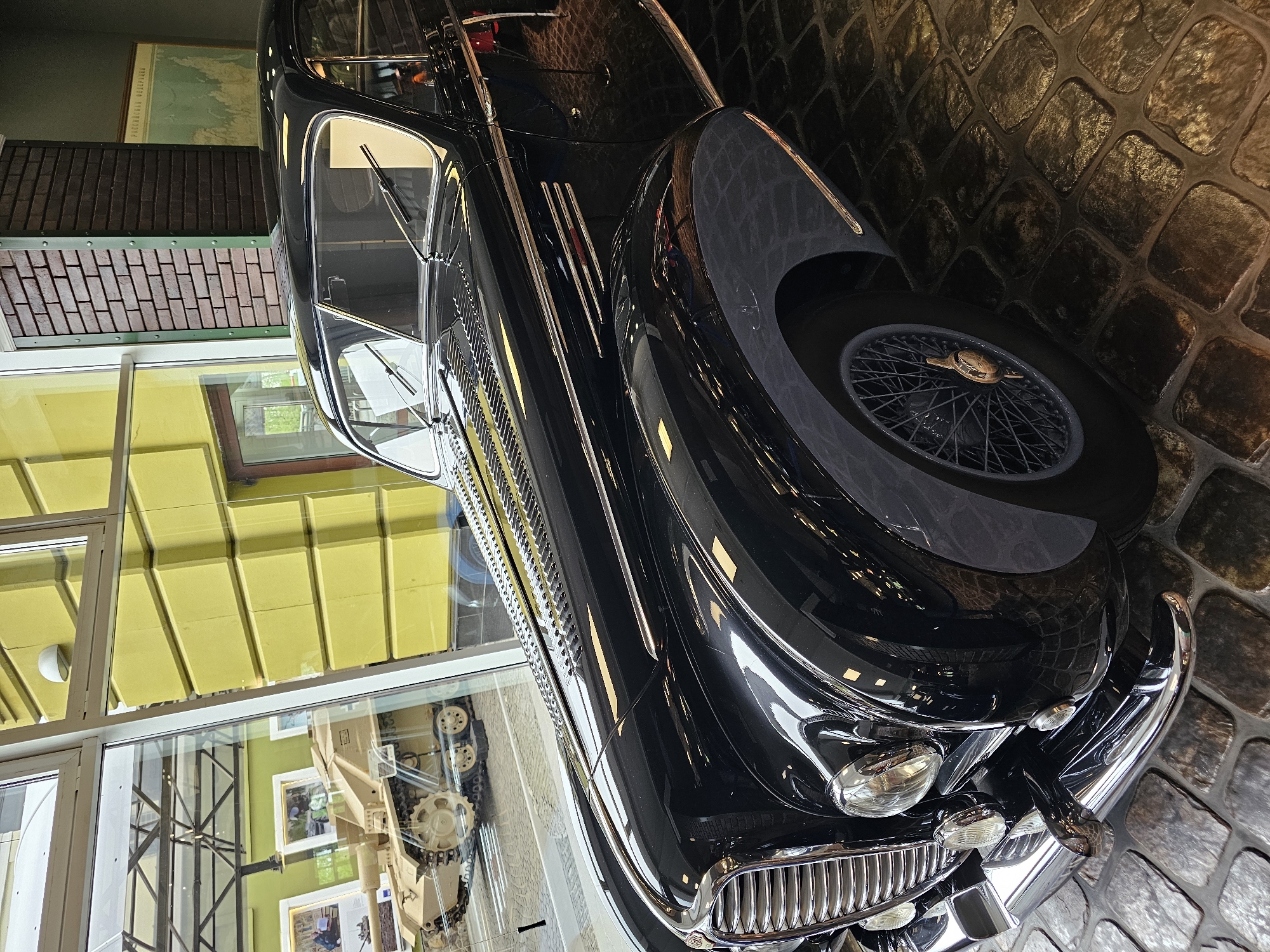 A black automobile, possibly a vintage model, is on display, likely indoors, as evidenced by the cobblestone floor and the reflection in its shiny surface. The image is slightly tilted, showing the front of the car with a large, curved fender, a spoked wheel, and chrome detailing on the grille and bumper. A display case with museum exhibits is visible through a window behind the car, along with other objects and photographs. The car's location is in Архангельское, Russia.