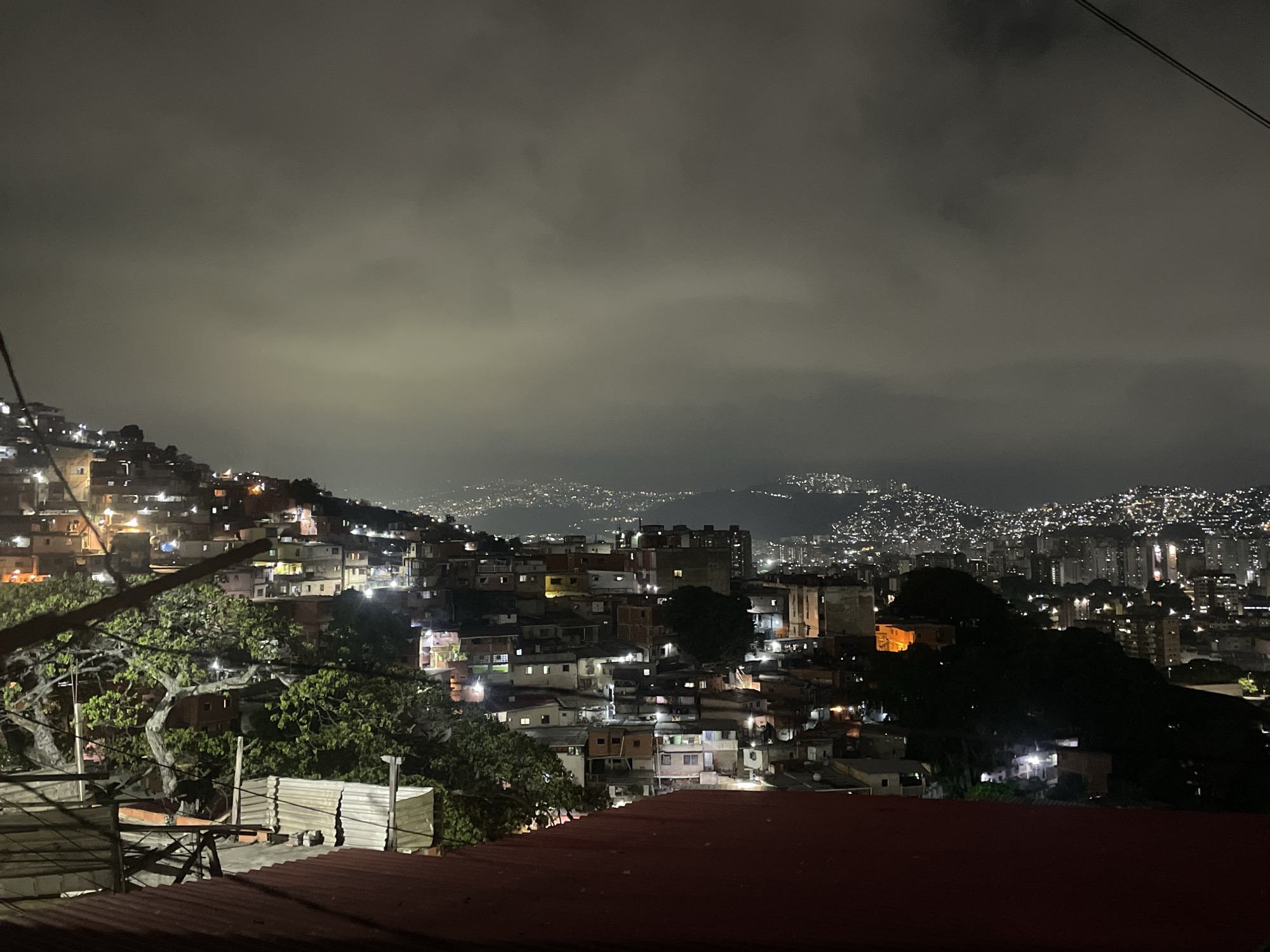The image displays a nighttime panoramic view of a densely developed hillside in Caracas, Venezuela, characteristic of an informal urban settlement (barrio), with the wider city extending into the background.
In the immediate foreground, the edge of a reddish-brown corrugated metal roof is visible at the bottom. Above this, a segment of a weathered, vertical corrugated metal fence and parts of a wooden structure are present on the left. The midground is dominated by a multitude of multi-story, irregularly constructed residential buildings, densely clustered along the hillside, with numerous windows emitting artificial light. Several large trees with green foliage are interspersed among these structures, and dark utility lines traverse the upper left. The background reveals a vast expanse of urban lights, stretching across distant hills and valleys, indicating the extensive cityscape of Caracas, all under a dark, heavily clouded sky. No human subjects or discernible actions are visible within the frame.