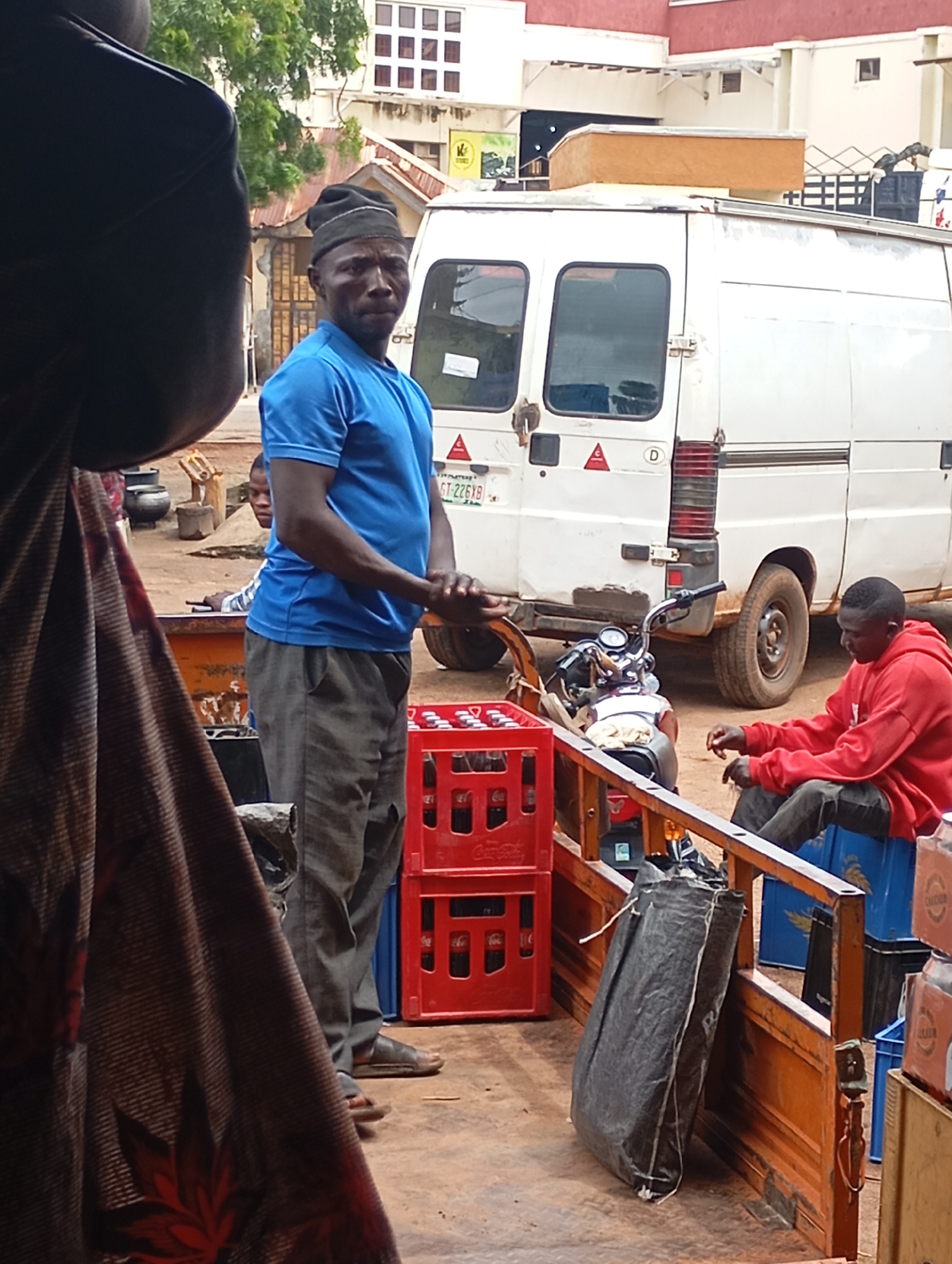 A man wearing a blue t-shirt and grey pants, with a black beanie on his head, stands in the back of an orange truck. He is facing towards the right of the frame, his arms crossed. To his right, a motorcycle is parked. Further to the right, another man in a red hooded sweatshirt is sitting on blue crates, also facing towards the right. In the background, a white van is parked behind the truck. There are stacks of red crates, filled with bottles, in the truck bed. A large black bag is leaning against the side of the truck. The scene appears to be outdoors, with a dusty ground and buildings visible in the background, suggesting a market or roadside setting. This image was taken in Shendam, Nigeria.