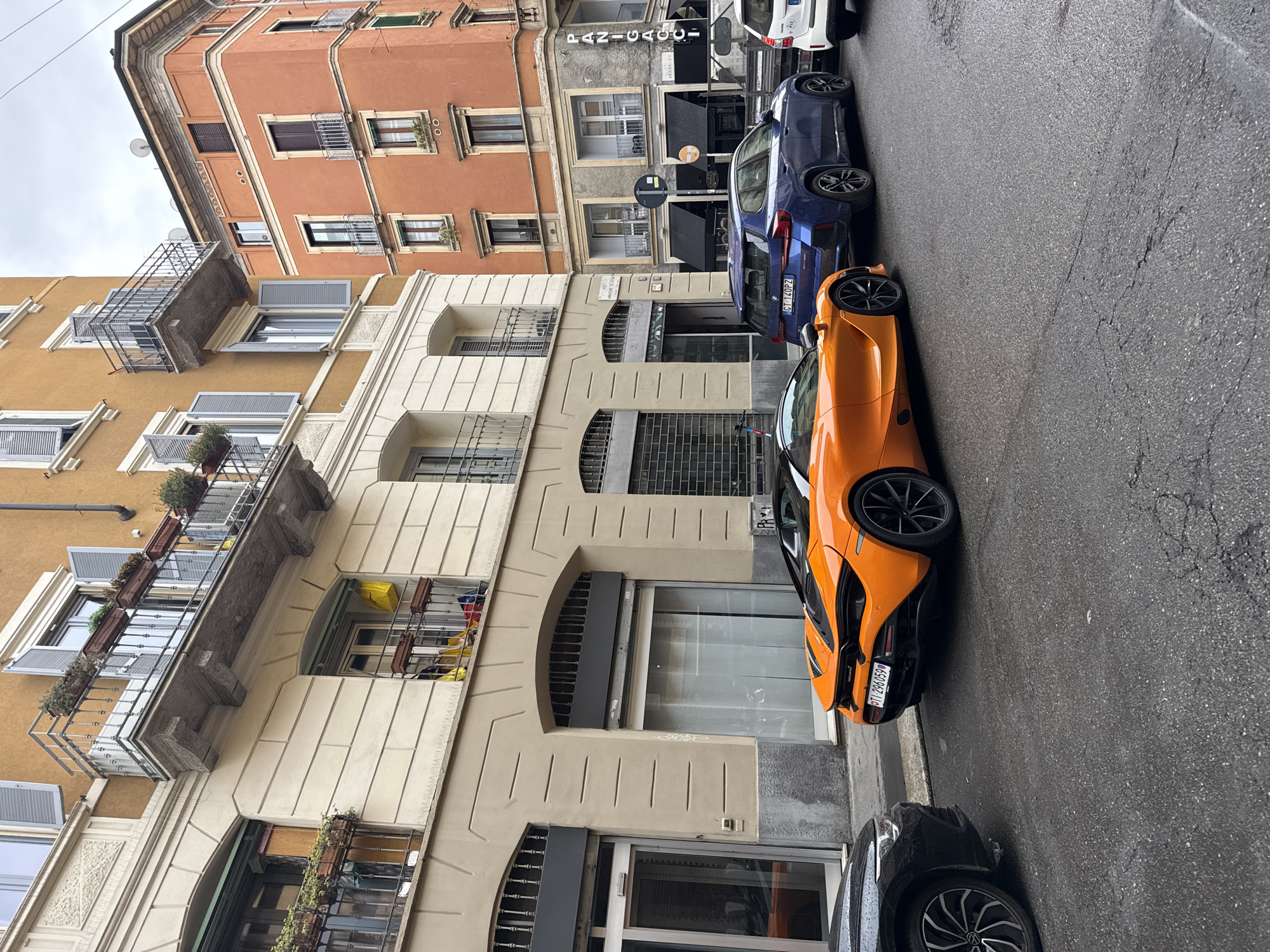 The image shows a street in Milan, Italy with several parked cars. The buildings are typical European-style with stucco facades, balconies, and window boxes. The parked cars include a bright orange McLaren, a dark blue BMW, and other vehicles that are partially visible. There are no people visible in the image. A sign for "PANICAGGI" can be seen on one of the buildings. The street is paved and slightly wet. The license plate on the orange McLaren is "FT 296 OS".