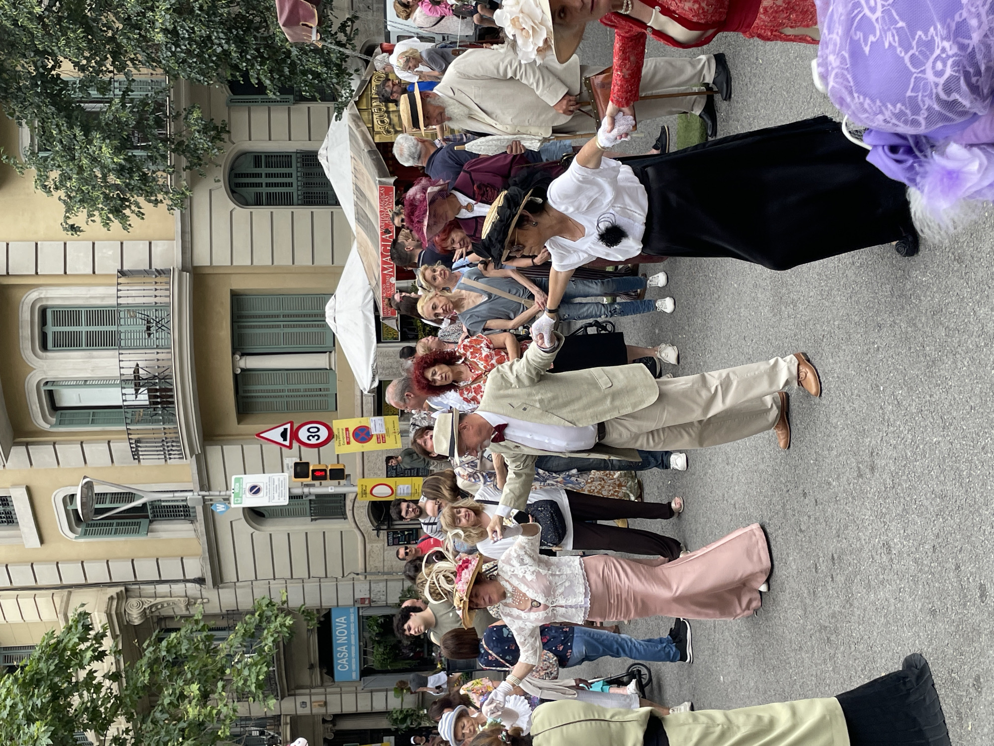 A street scene in Barcelona, Spain, shows a crowd of people dressed in late 19th-century and early 20th-century clothing, suggesting a historical recreation or themed event. The people, mostly in pairs, are holding hands, moving as if they are performing a procession or dance.
Many are wearing elaborate hats and period-appropriate attire, including long dresses, suits, and gloves. The background features buildings with architectural details typical of Barcelona, including balconies and window shutters.
Street signs and a traffic light are visible. One sign reads "CASA NOVA."