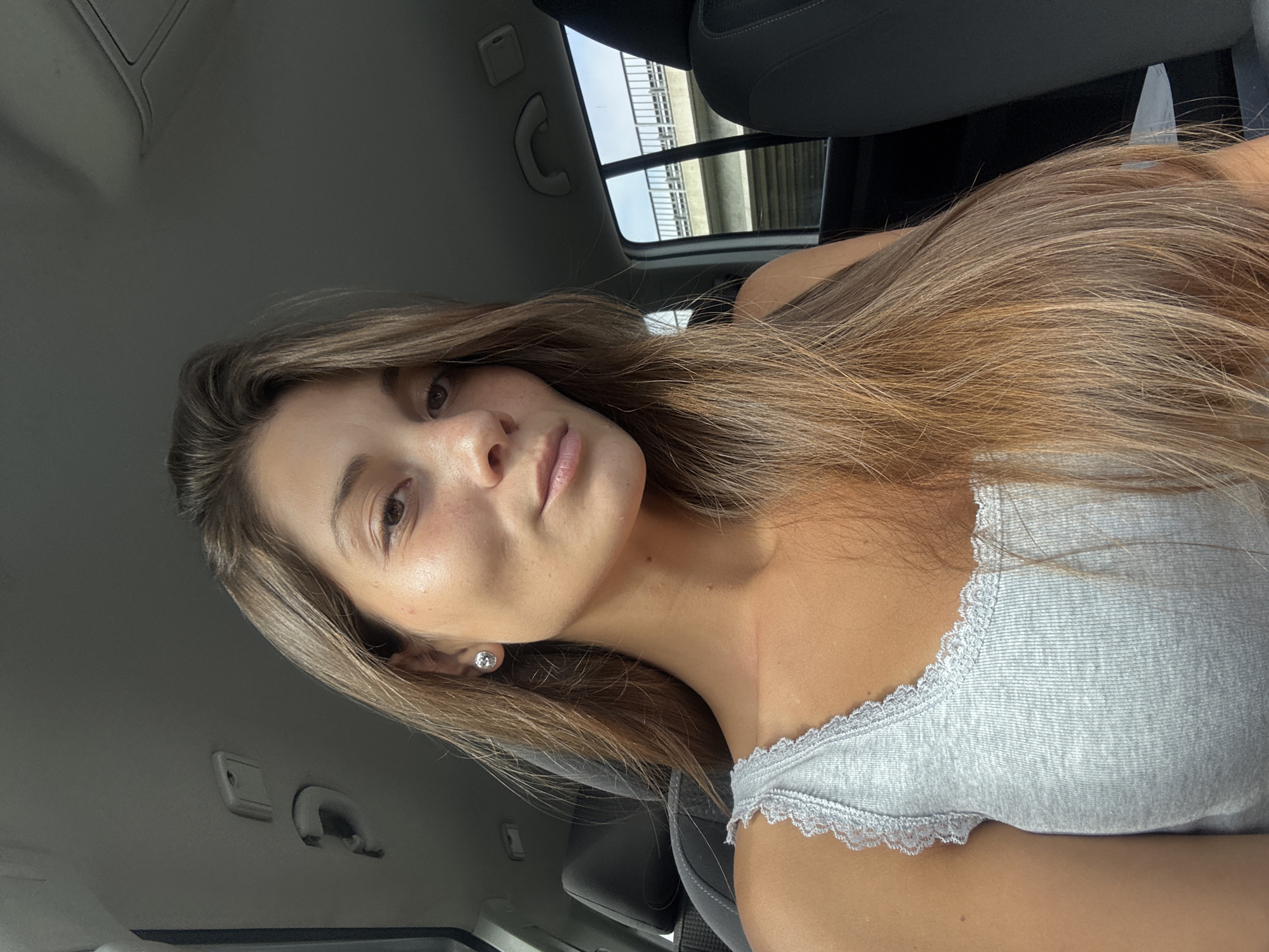 A light-skinned woman with long brown hair is pictured inside a car. She wears a gray tank top with lace trim. She is possibly on a bridge. The location cannot be determined to be Burgas, Bulgaria.