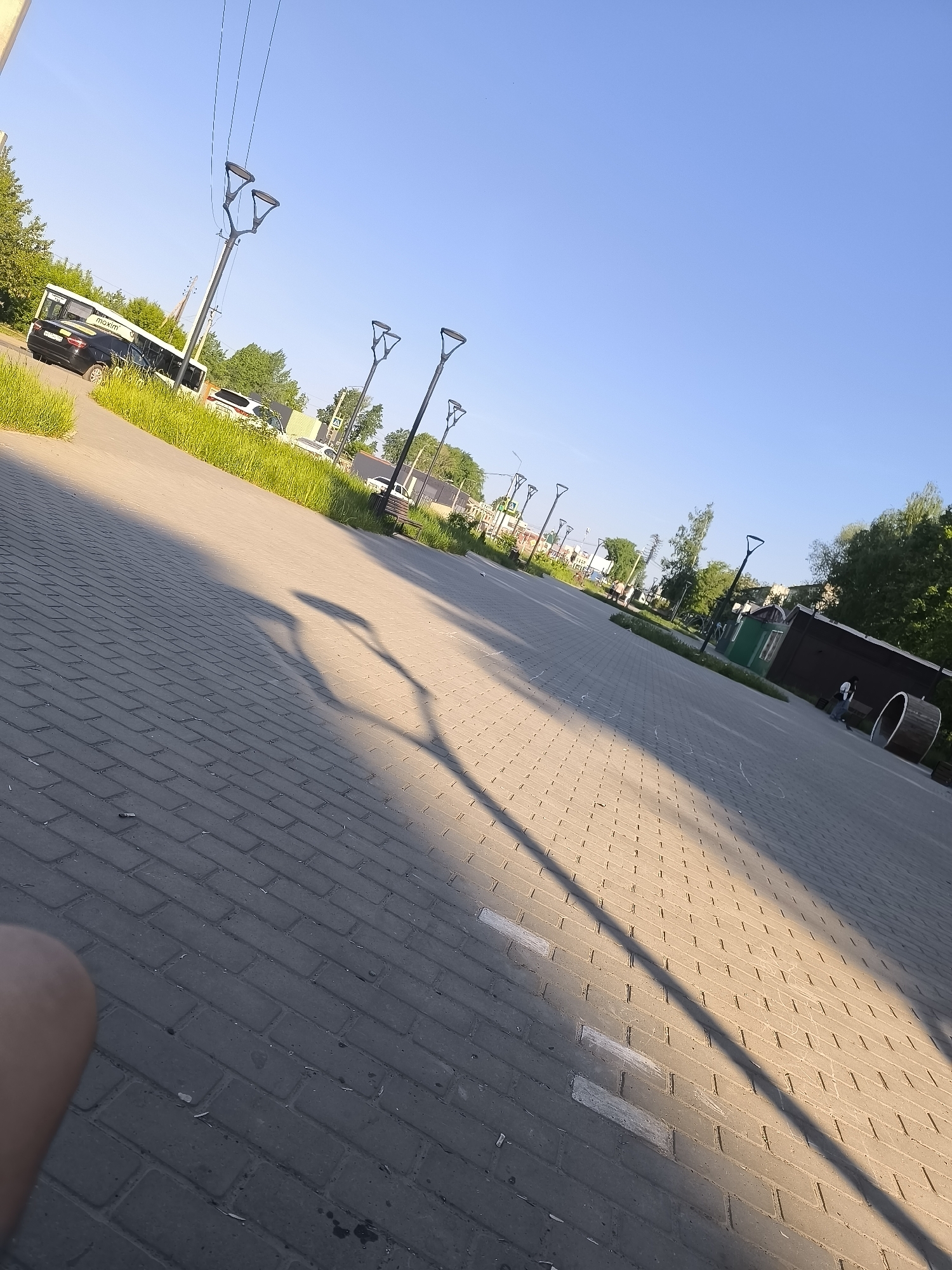 The image shows a paved pathway in Unknown, Unknown, stretching into the distance with multiple streetlights lining the side. A long shadow of a streetlight stretches across the pathway. In the background, there is a bus and several other vehicles parked along the edge of the pathway. Vegetation, including trees and grass, is visible along the roadside and in the distance. A blue sky is present in the background. A portion of a person's leg is visible in the bottom left corner of the image. A small, enclosed structure, possibly a shed or utility building, is visible on the right side. A cylindrical object is also present on the right side.