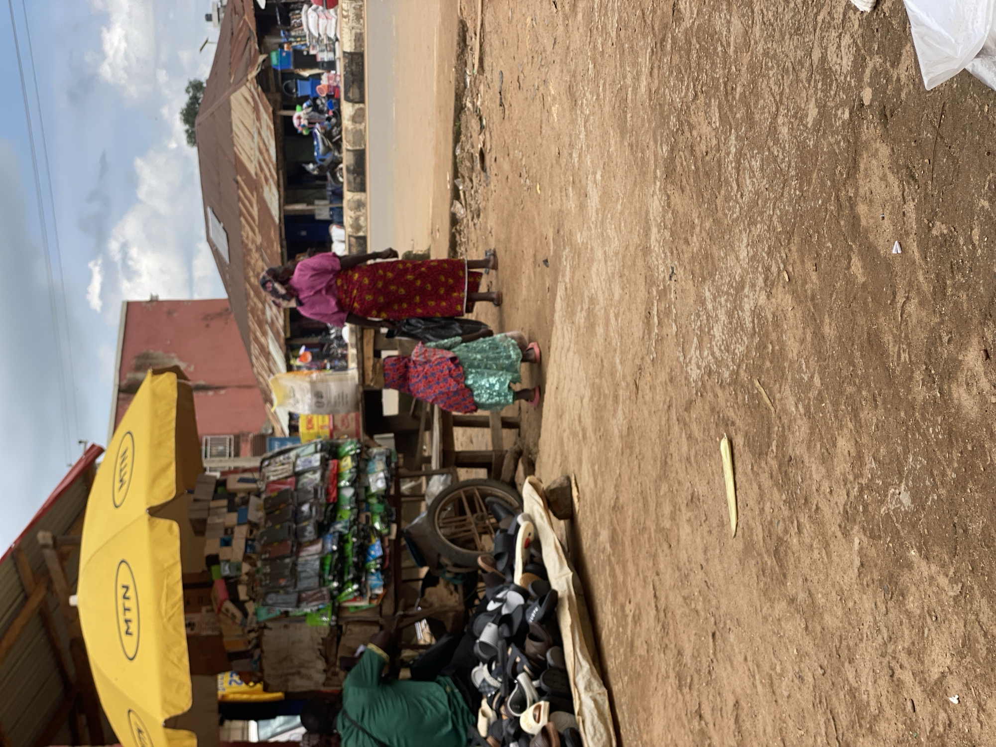 This is an outdoor, ground-level, eye-level shot taken in Kafanchan B, Nigeria. A vendor stands behind a stall topped with a yellow MTN-branded umbrella. The stall displays a variety of items, including numerous phone cases and accessories on the left and a pile of footwear on the left foreground. To the right of the stall, a woman wearing a pink top and a patterned skirt walks away from the camera, holding a black plastic bag. Beside her, a young child wearing a green dress and headscarf walks in the same direction. In the background, a street is visible with buildings and parked motorcycles. The ground is dirt, and the sky is overcast.