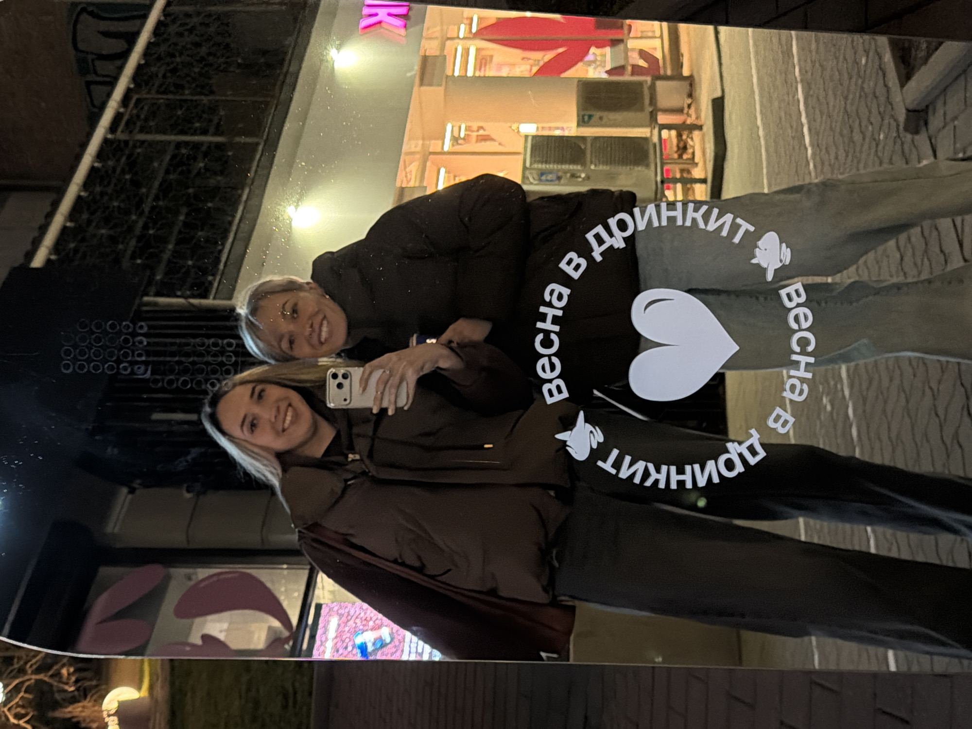 The image displays two adult females posing for a mirror selfie at night on a paved urban street in Rostov-on-Don, Russia. The specific location is identified as a "Drinkit" establishment, with the text "Весна в Дринкit" (Spring in Drinkit) and a partial "принкит" (prinkit) visible on the reflective surface, along with pink neon signage on the building facade in the background.
The female on the left has long blonde hair, wears a dark brown puffer vest over a dark long-sleeved top, dark trousers, and carries a dark shoulder bag on her left side. The female on the right has short blonde hair and is wearing a dark puffer jacket and light-colored jeans. She is holding a light-colored smartphone with both hands at chest level, capturing the reflection. Both individuals are smiling and looking directly towards the camera's perspective.
The reflective surface, possibly an outdoor mirror, also features a prominent white heart graphic. The background depicts the brightly illuminated exterior of a commercial building, characteristic of the "Drinkit" brand, with large windows, multiple external air conditioning units, and a visible railing on an upper level. To the far left of the frame, a segment of a park-like area with grass, tree branches, and an illuminated spherical light fixture is visible. The ground is paved with dark grey interlocking pavers.