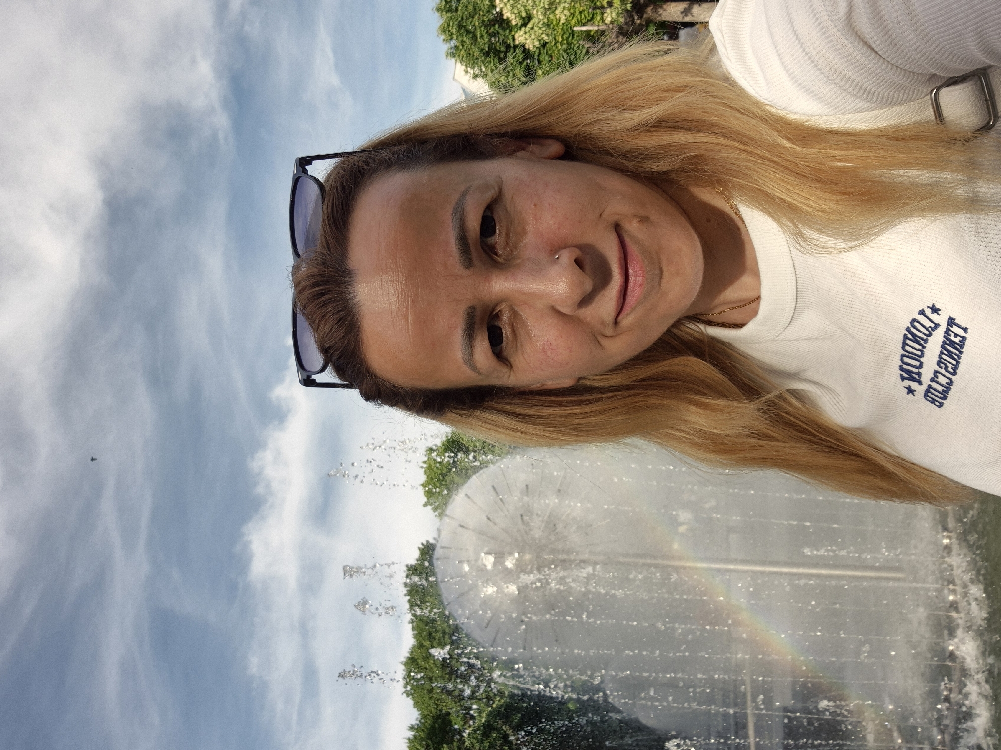 A woman with long, blonde hair and sunglasses perched on her head takes a selfie in front of a modern, spherical fountain that is spraying water, creating a rainbow. She is wearing a white t-shirt with blue lettering that appears to say "LONDON TENNIS CLUB." The background shows green trees and a cloudy blue sky.