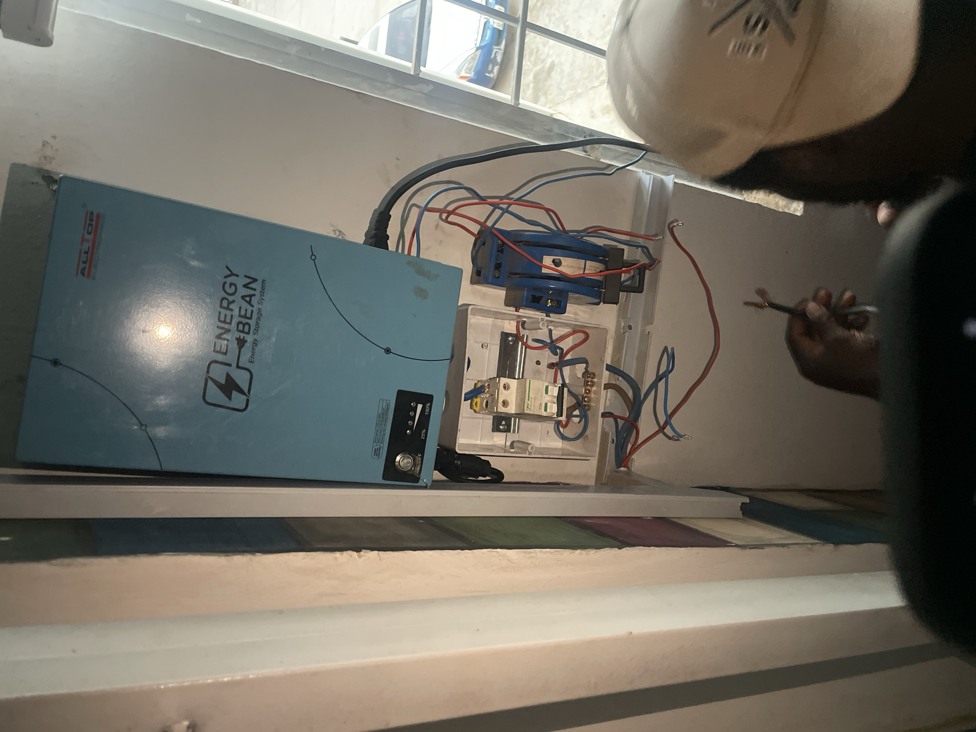 The image shows an electrician working on an "Energy Bean" energy storage system inside a room. The system is mounted in a white electrical box, with visible red and blue wiring connected to a blue electrical component and a smaller, white component. The electrician, wearing a white cap, is holding pliers and working on the wiring. The scene appears to be indoors with a window visible in the background. The location is Hayin Banki, Nigeria, as specified.