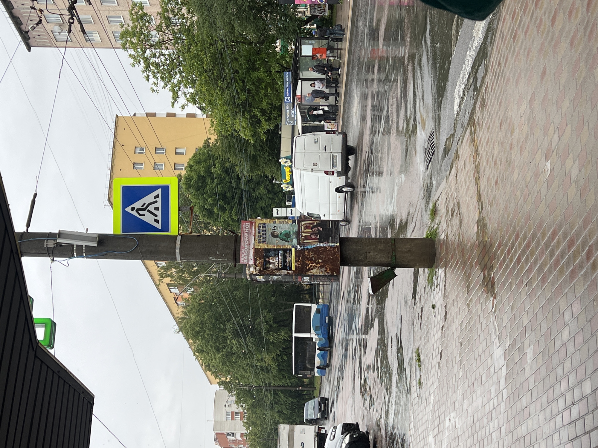 This image captures a wet urban street scene in Ternopil, Ukraine, likely after a recent rain shower, given the glistening wet pavement and overcast sky. The photograph is taken from a slightly elevated position, looking down the street.
**Subjects and Scene:** The foreground features a tiled sidewalk and the edge of a building on the left. The main street is visible, with dark, reflective asphalt covered in puddles and runoff. A prominent lamppost stands near the center-left, adorned with a blue and yellow pedestrian crossing sign. Several utility wires crisscross the sky. On the right side of the street, there's a bus stop with a shelter, where several individuals are seated or standing, waiting. Beyond the bus stop, lush green trees line the street, partially obscuring multi-story residential buildings in the background, one with a distinct yellow facade.
**Activity and Objects:** People are seen waiting at the bus stop. Various vehicles are present on the wet road, including a white delivery van parked near the bus stop, a blue and white bus further down the street, and other cars. The white van's rear is visible, showing a European-style license plate. Posters and advertisements are plastered on the lamppost, some appearing worn or rusty. A storm drain grate is visible in the wet pavement.
**Notable Details:** The wet conditions are a key feature, with reflections off the puddles and a general somber atmosphere due to the grey sky. The time of day appears to be daylight.
**Visible Text:**
* **Pedestrian Sign:** A standard blue square sign with a yellow border, depicting a black walking figure on a white triangle, indicating a pedestrian crossing.
* **Bus Stop Sign:** On the bus stop shelter, Cyrillic text is visible, including "ТЕРНОПІЛЬСЬКОЇ МІСЬКОЇ РАДИ" (Ternopil City Council) and "ЗУПИНКА" (Stop), confirming the location context.
* **Posters on Pole:** Various advertisements are posted. Fragments like "ЗАБОРОНЕНО!" (Forbidden!), "СТУЛІТЬ" (Close/Shut), and "ГОЛОВКИ" (Heads - likely part of a larger phrase) are discernible. One poster appears to be for a movie or event, showing faces.