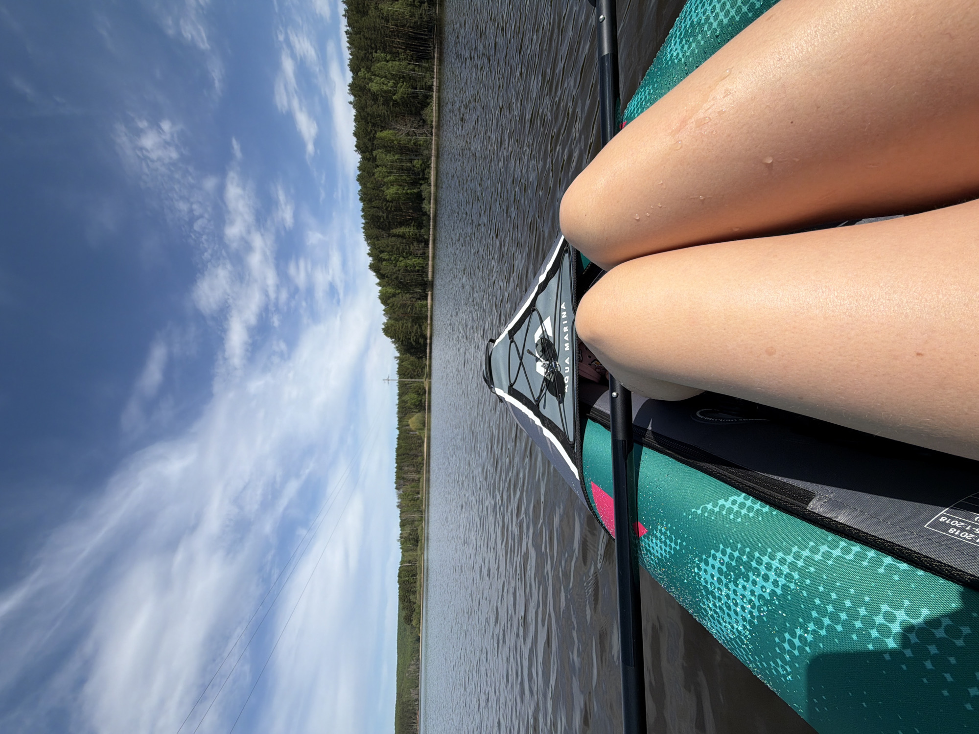 The image shows a first-person view from inside a kayak on a body of water. The legs of a person are visible in the foreground. The kayak is green and gray and labeled "AQUA MARINA." A paddle is positioned on the right side of the kayak. In the background, there is a calm body of water, a line of trees, and a blue sky with scattered clouds. Power lines are visible in the background. The location is Chaykovsky, Russia.
