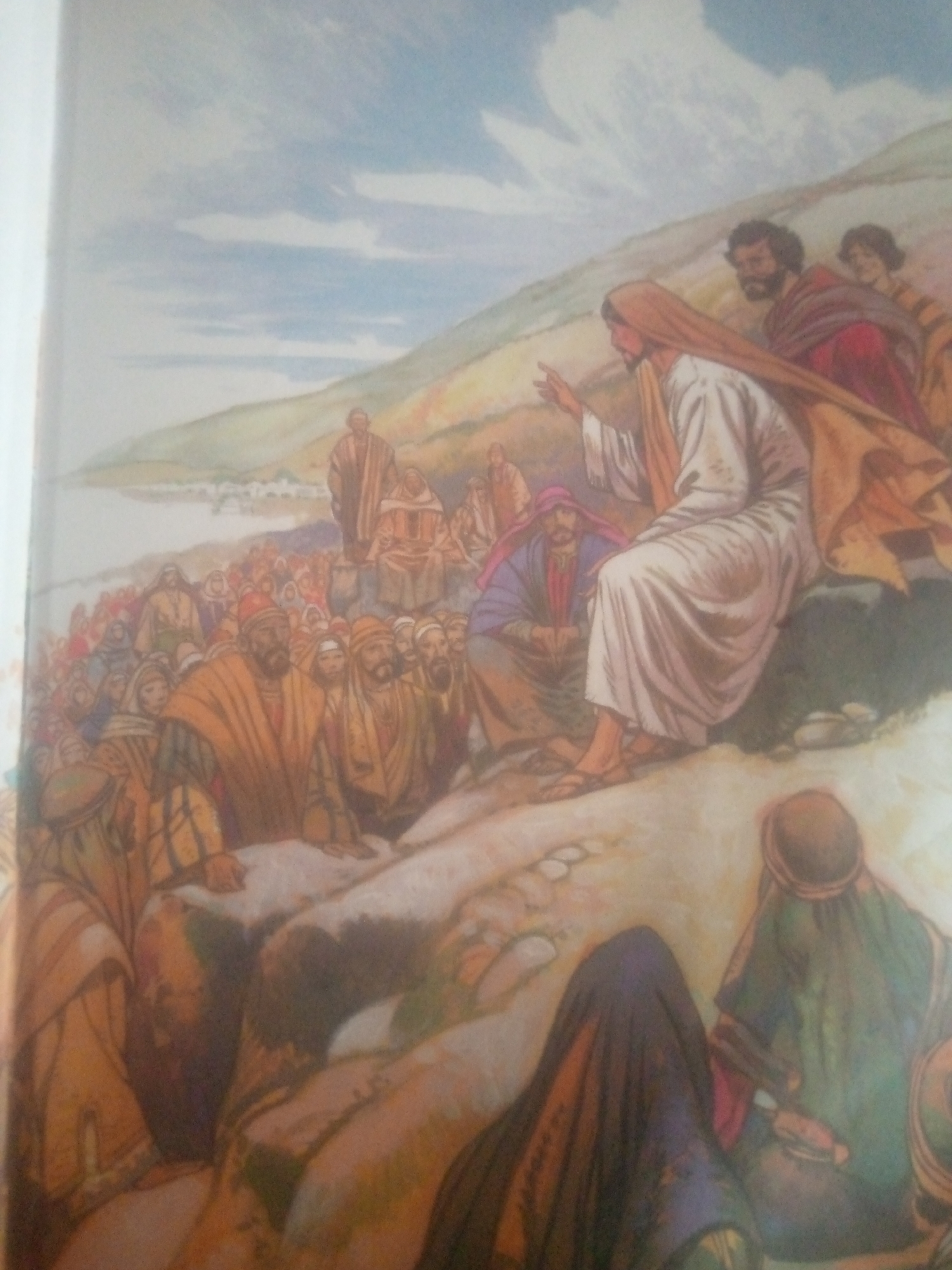 The image depicts a religious scene, likely an illustration from a religious text, featuring Jesus speaking to a crowd of people. Jesus, clad in white robes and a tan head covering, is seated on a rocky outcrop, gesturing with his right hand. He is surrounded by a large gathering of individuals, many of whom are also wearing robes and head coverings in earthy tones. Some figures are seated, while others are standing, all facing towards Jesus. The setting appears to be outdoors, with a hillside and a body of water in the background, suggesting a gathering in a natural landscape. The sky above is blue with white clouds. There is no information in the image that would indicate the location is the city of Jauro Yinu, Nigeria.
