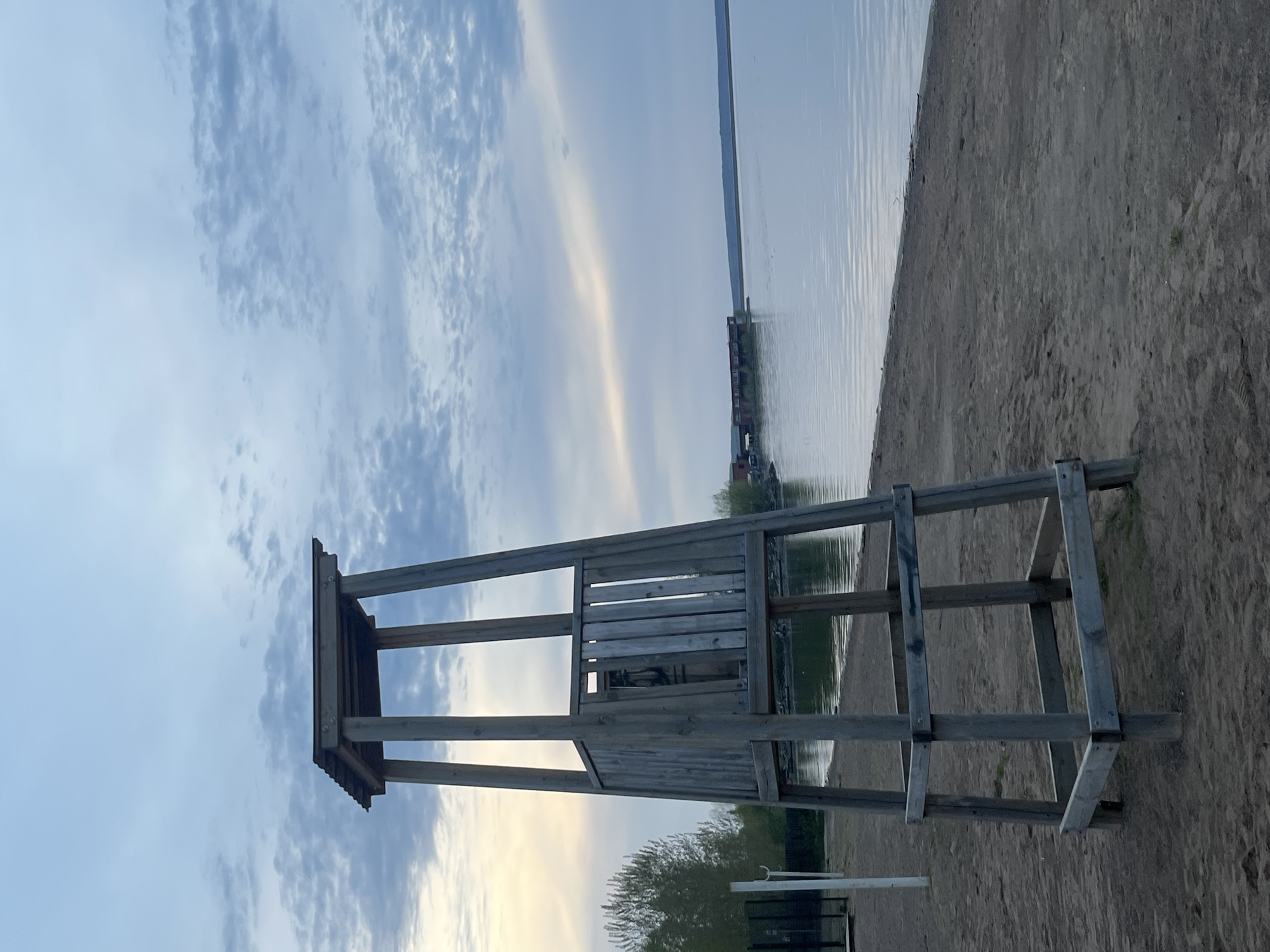 The image shows a lifeguard tower on a sandy beach. The tower is made of weathered wood and consists of a raised platform with a roof, supported by a wooden frame. The beach borders a body of water. In the background, across the water, are buildings and structures. The sky is partly cloudy. The location is identifiable as the city of Chaikovsky, Russia.