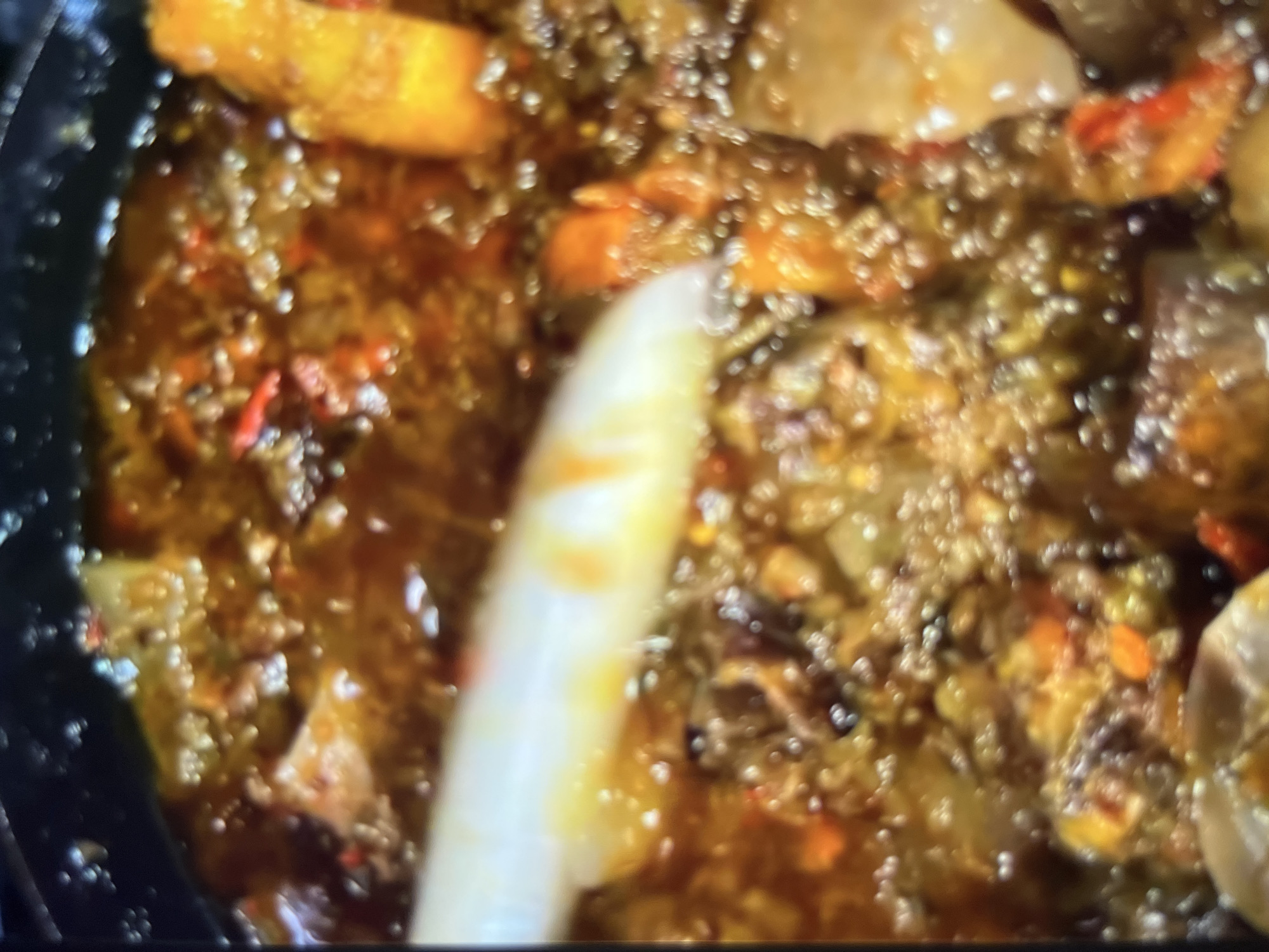 The image is a close-up, slightly blurry shot of a pot of food being cooked. The food appears to be a stew or soup with chunks of meat or vegetables in a rich, dark brown sauce. There are visible red specks, likely from chili peppers, and some lighter colored pieces which could be potatoes or carrots. Bubbles on the surface indicate it is actively cooking. The food is in a dark-colored pot, the rim of which is visible at the top and left of the frame. A white utensil is partially visible in the upper left quadrant, suggesting it is being stirred or served. There are no discernible landmarks or contextual clues within the image to suggest a specific location in Abuja, Nigeria. The scene is limited to the contents of the pot.
