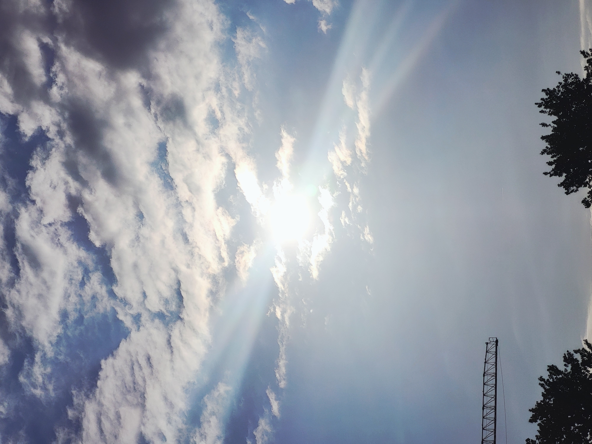 A crane extends horizontally across the upper left portion of the frame against a hazy sky with sunbeams. The sky is a mix of bright blue and white and grey clouds, with the sun prominently visible and radiating light in streaks. Silhouetted trees are visible at the bottom of the image.