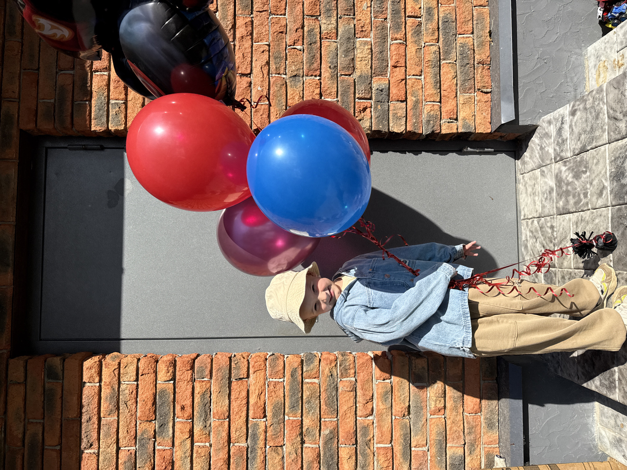 A young child wearing a light-colored bucket hat and a denim jacket stands in front of a brick wall and a gray door, holding a bouquet of red, pink, and blue balloons. The child is smiling and looking at the camera. The balloons are tied with red ribbons. The ground is paved with gray tiles. The scene appears to be outdoors, possibly in a residential area. The location is Chaikovsky, Russia.
