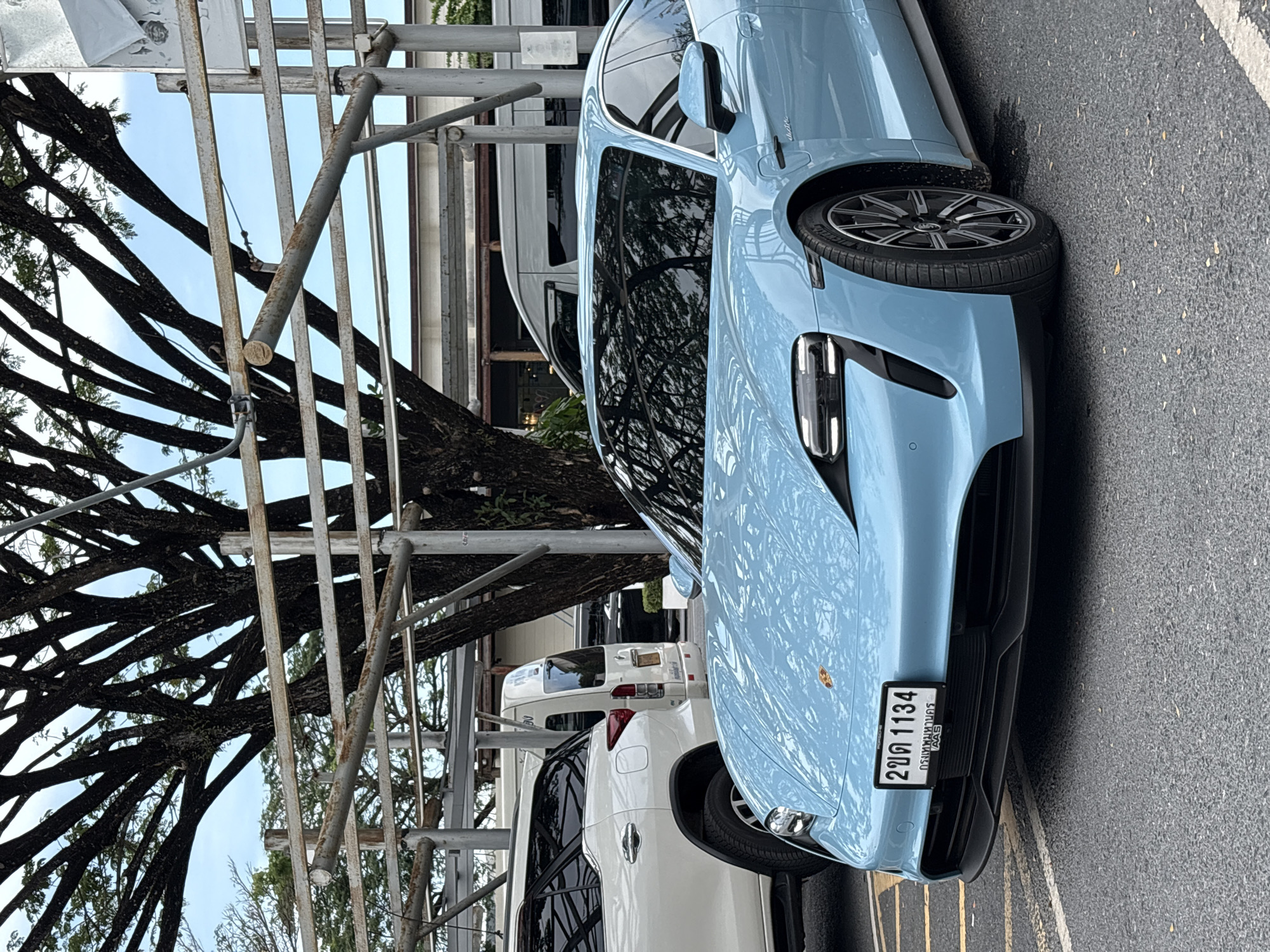 In Choeng Thale, Thailand, a light blue Porsche Taycan stands parked beside scaffolding. A white SUV and camper are parked behind it. A large tree with bare branches is situated next to the scaffolding. The car's license plate is visible. The ground is asphalt.