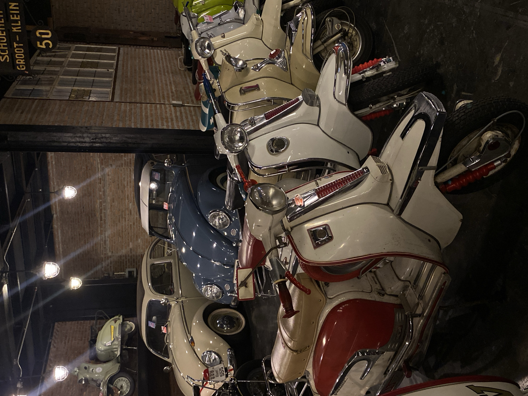 The image depicts a collection of vintage scooters and cars stacked closely together. The color palette is dominated by off-white, red, blue, and green hues. The vehicles include scooters with distinctive chrome accents and rounded car models reminiscent of vintage Volkswagen Beetles. Overhead, visible light fixtures illuminate a brick wall and a sign reading "SCHUENELI, GROOT - KLEIN, 50". The location is not specified in the provided image details or hints.