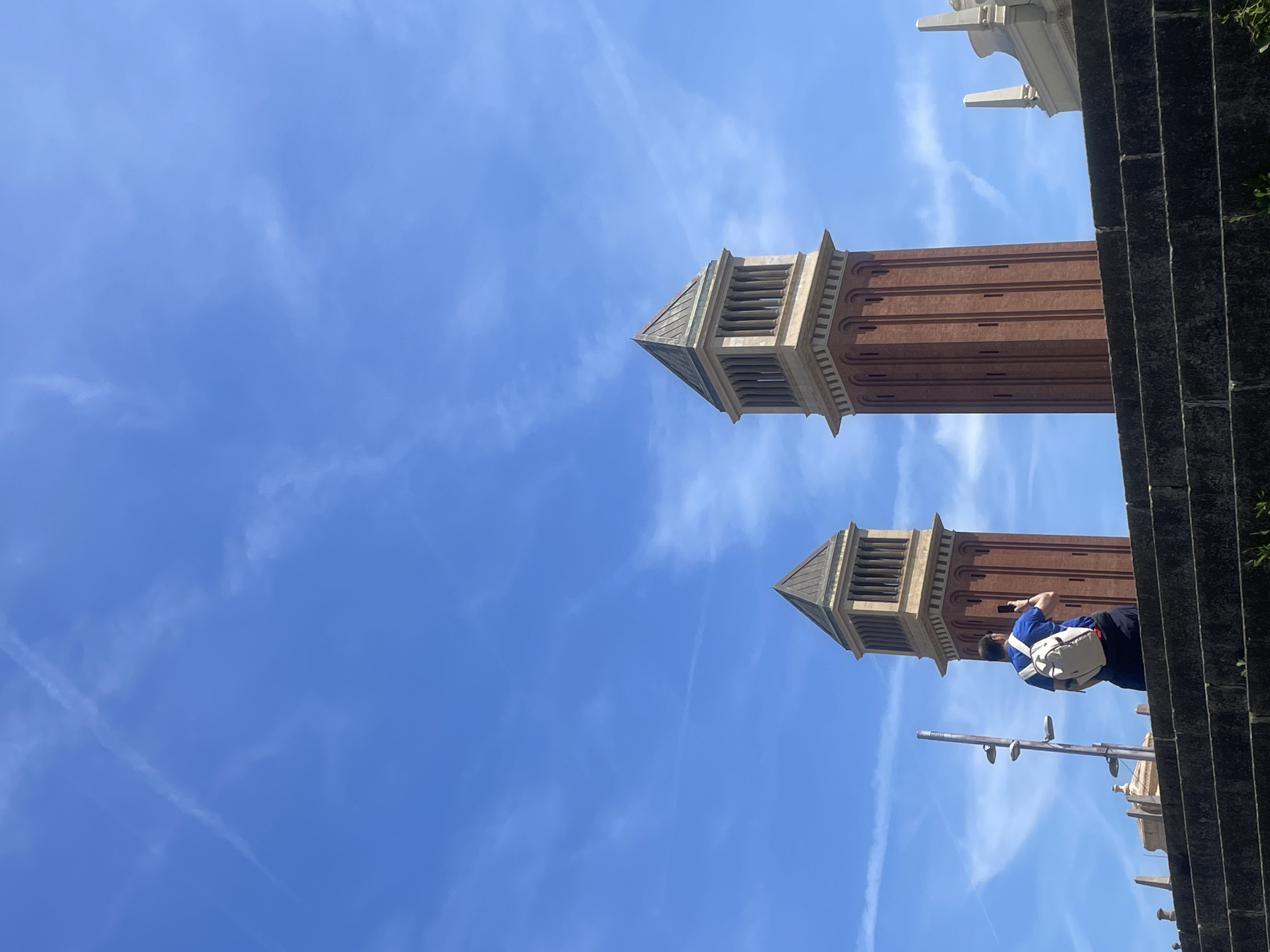 The image displays an outdoor architectural scene in Barcelona, Spain, centered around two prominent, identical towers set against a bright blue sky. The towers are tall, with bodies constructed from reddish-brown brick. Their upper sections feature lighter-colored stone or concrete elements, including slatted openings suggestive of belfries or observation points, topped with dark, pyramidal roofs.
In the foreground, slightly to the right, a person is visible, wearing a blue short-sleeved shirt and dark pants, with a light-colored fanny pack or small bag slung across their chest. They are holding a mobile phone, seemingly capturing the view of the towers. Parts of dark stone steps and a metal railing or light pole are also present in the lower right and mid-ground.
The sky is clear and blue, with several wispy white clouds and faint airplane contrails streaking across it, indicating a sunny day. The bright lighting casts subtle shadows on the structures. These twin towers are characteristic of the Venetian Towers at Plaça d'Espanya in Barcelona. No visible text is present in the image.