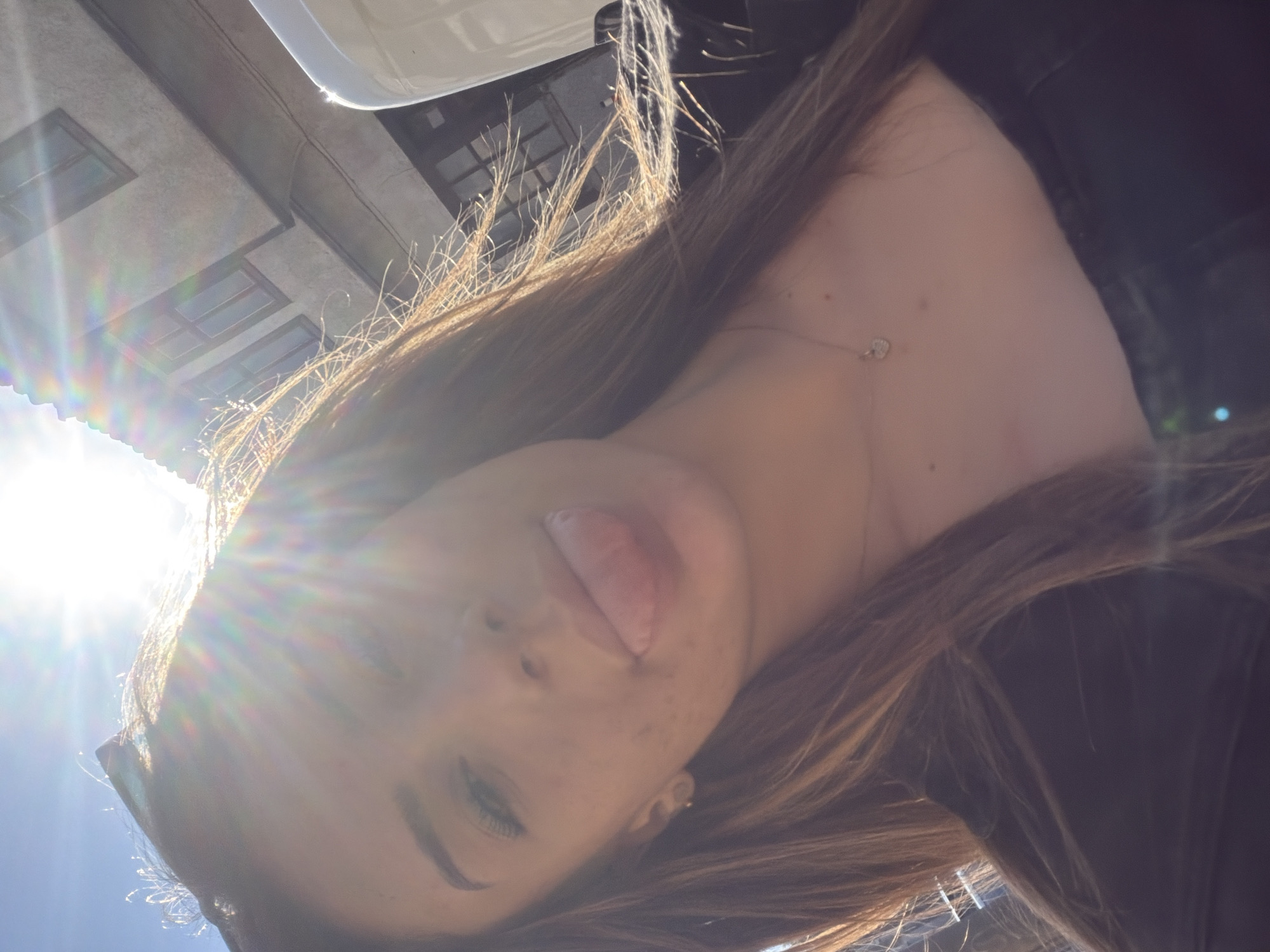 A young woman with long, brown hair, wearing a dark top and a heart-shaped pendant necklace, sticks her tongue out at the camera in a selfie. The sun is very bright, creating lens flare across the image and obscuring parts of the background, which appears to be an urban setting with a building and a white vehicle. The woman has light skin with some freckles and appears to be outdoors. The scene is likely set in Shumen, Bulgaria, given the context.