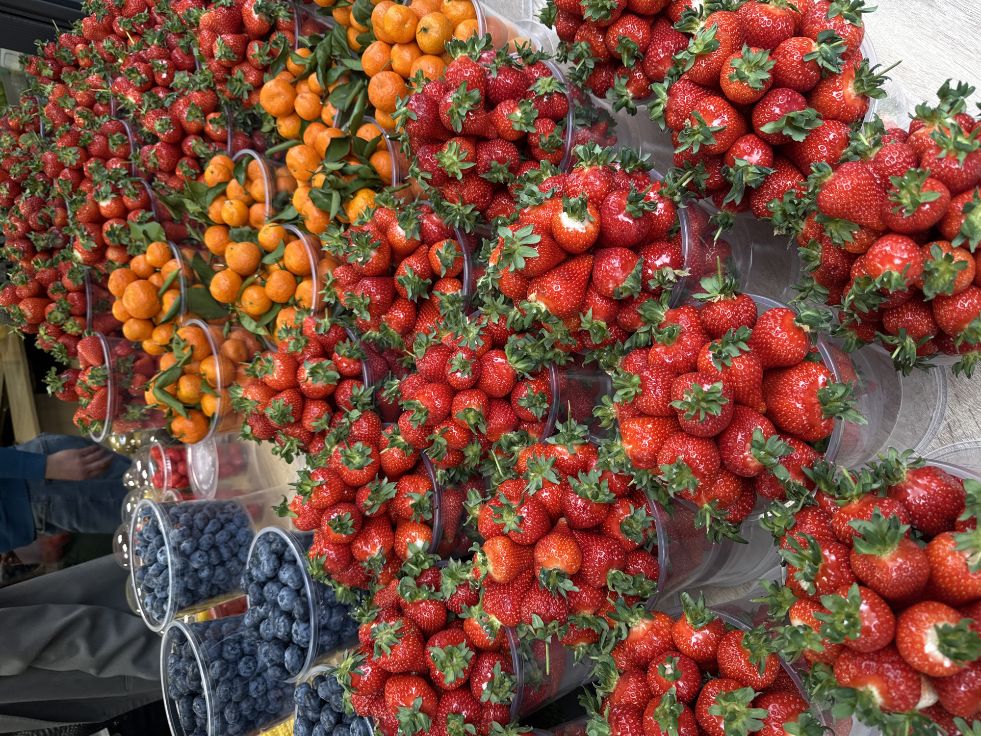 This image captures a vibrant and abundant display of fresh fruit, likely at a market stall or grocery store in Yerevan, Armenia. The scene is dominated by numerous clear plastic containers, each brimming with ripe produce.
The most prominent fruit visible is bright red strawberries, presented in rows of transparent containers that stretch across the frame, particularly on the right and extending towards the center and left. Many strawberries still have their green calyxes attached, indicating freshness. Interspersed among the strawberries are containers filled with smaller, orange-colored citrus fruits, likely tangerines or mandarins, some still bearing small green leaves on their stems, adding a touch of natural verdancy to the display. Towards the lower-left corner, several clear plastic cups are stacked and filled with dark blue blueberries, contributing another burst of color to the arrangement.
The fruits are neatly organized, suggesting a retail setting where they are prepared for sale. A person's legs, clad in blue jeans and grey trousers, are partially visible in the lower-left section of the image, hinting at a shopper or vendor present at the display. The overall lighting is bright and even, indicating either indoor artificial lighting or a well-lit outdoor market space during the daytime. There is no visible text or branding within the frame. The image conveys a sense of freshness and variety, showcasing plentiful seasonal offerings.
