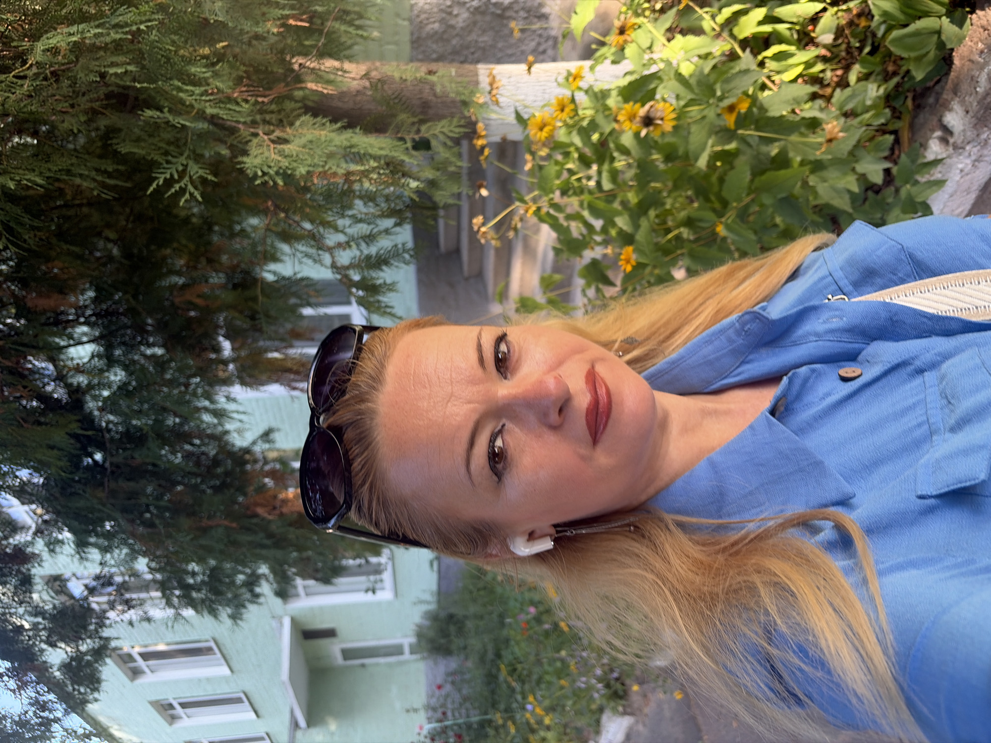 An outdoor medium close-up selfie shows a fair-skinned blonde woman with sunglasses perched on her head, white earbuds in her ears, and wearing a blue collared shirt. She has winged eyeliner and dark lipstick. Behind her, a light green building with white-framed windows and a small balcony is visible, as is dense green foliage with yellow flowers and a tree. The background suggests a residential area, possibly a park or garden. The lighting indicates it is daytime.