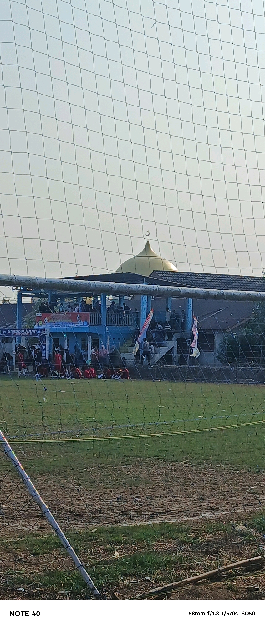 The image shows a view of a soccer field in Simbur Naik, Indonesia, through a net. In the background, there is a blue building with a balcony or seating area where spectators are gathered. A golden dome with a crescent moon atop it, indicative of a mosque, is visible above the building. Several people, some wearing red jerseys, are on the field, with some sitting on the grass. The field itself appears to be a mix of green grass and dry, patchy ground. The sky is bright and hazy. The text "NOTE 40" and "58mm f/1.8 1/570s ISO50" are present at the bottom of the image, likely indicating the photo's origin or settings.