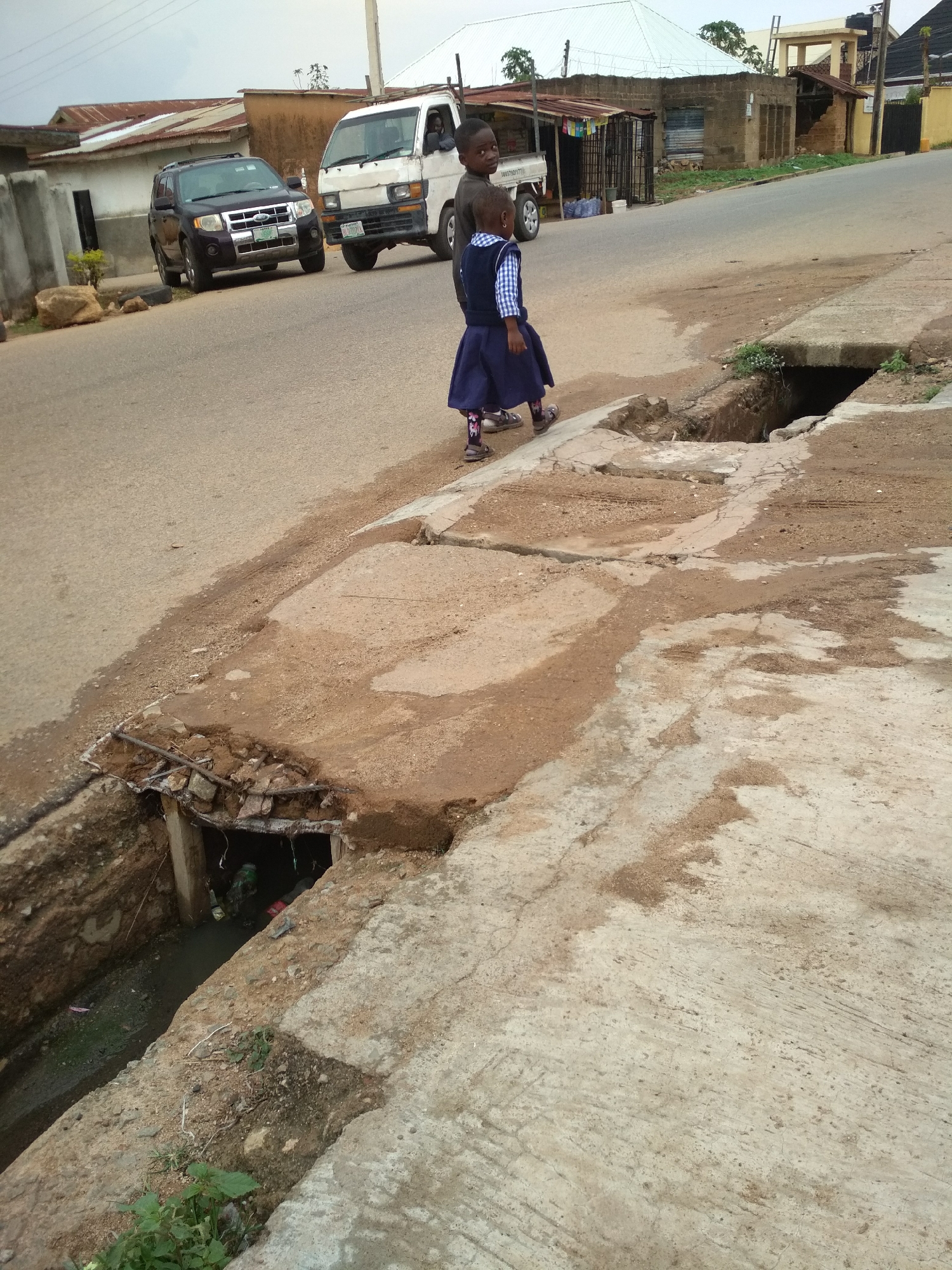 The image depicts an outdoor scene in what appears to be an urban or semi-urban environment in Jos, Nigeria. Two young children are prominent in the foreground, walking along the side of a paved road. The child closer to the camera is a young girl, dressed in a dark blue skirt, a blue and white checkered shirt, and sandals over patterned socks, strongly suggesting a school uniform. She is followed by a slightly older boy wearing a dark top. Both children have their backs partially to the camera, looking towards the right side of the frame.
The road itself is paved but shows considerable wear and patches of dirt. A critical feature of the scene is the severely damaged concrete drainage system running parallel to the road. A large section of the drainage ditch in the foreground has collapsed, exposing rusty rebar and a visible channel containing stagnant water and various pieces of litter and debris. Further along the road, other sections of the drainage system also appear to be in disrepair.
In the middle ground, two vehicles are present on the road: a dark Ford Escape SUV and an older model white van or mini-truck. Behind these vehicles, and extending into the background, are various buildings, many featuring corrugated metal roofs, typical of residential or commercial structures. The sky is overcast, indicating a cloudy day with diffuse lighting. The overall impression is one of a community grappling with infrastructure challenges, particularly regarding its drainage and road conditions.
Visible text includes license plates on both the Ford Escape and the white van, though the full details are not clearly legible. A number "00717017" is visible on the side of the white van.