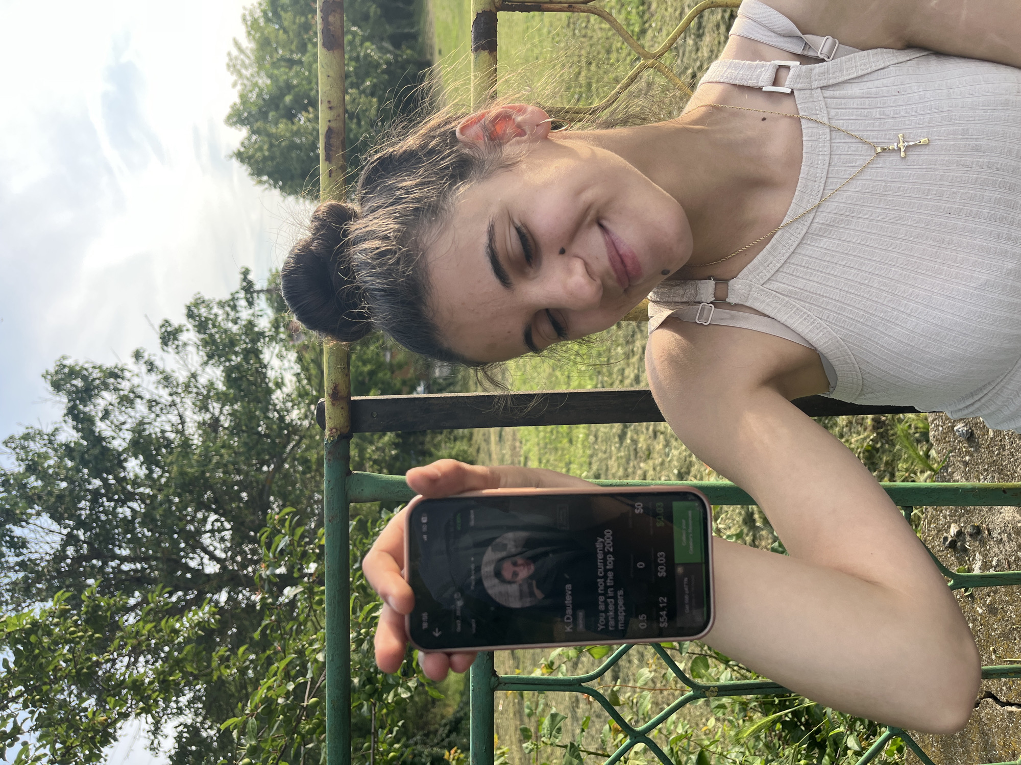 An outdoor portrait, taken in Lozarevo, Bulgaria, features a young woman with her head tilted to the left due to the image's clockwise rotation. She has dark hair tied in a bun, dark eyebrows, and a mole on her cheek. Her expression is a relaxed, soft smile with partially closed eyes, conveying a sense of ease or amusement. She wears a light-colored, ribbed tank top with prominent strap details and a gold chain necklace with a crucifix pendant resting on her chest.
In her right hand, the woman holds a smartphone in a pinkish case, displaying its screen directly towards the viewer. The screen shows what appears to be a profile within an application, featuring a circular profile image and the name "K.Dauteva." Text below reads: "You are not currently ranked in the top 2000 mappers." Further visible text includes numerical values: "0.5," "0," "$54.12," "$0.03," "$0," and "$0.03."
The setting is outdoors during the daytime, under a bright sky with some clouds. Behind the woman, a green and rusty metal fence is visible, alongside abundant green trees and foliage, indicating a natural or rural environment. The overall atmosphere is casual and relaxed.