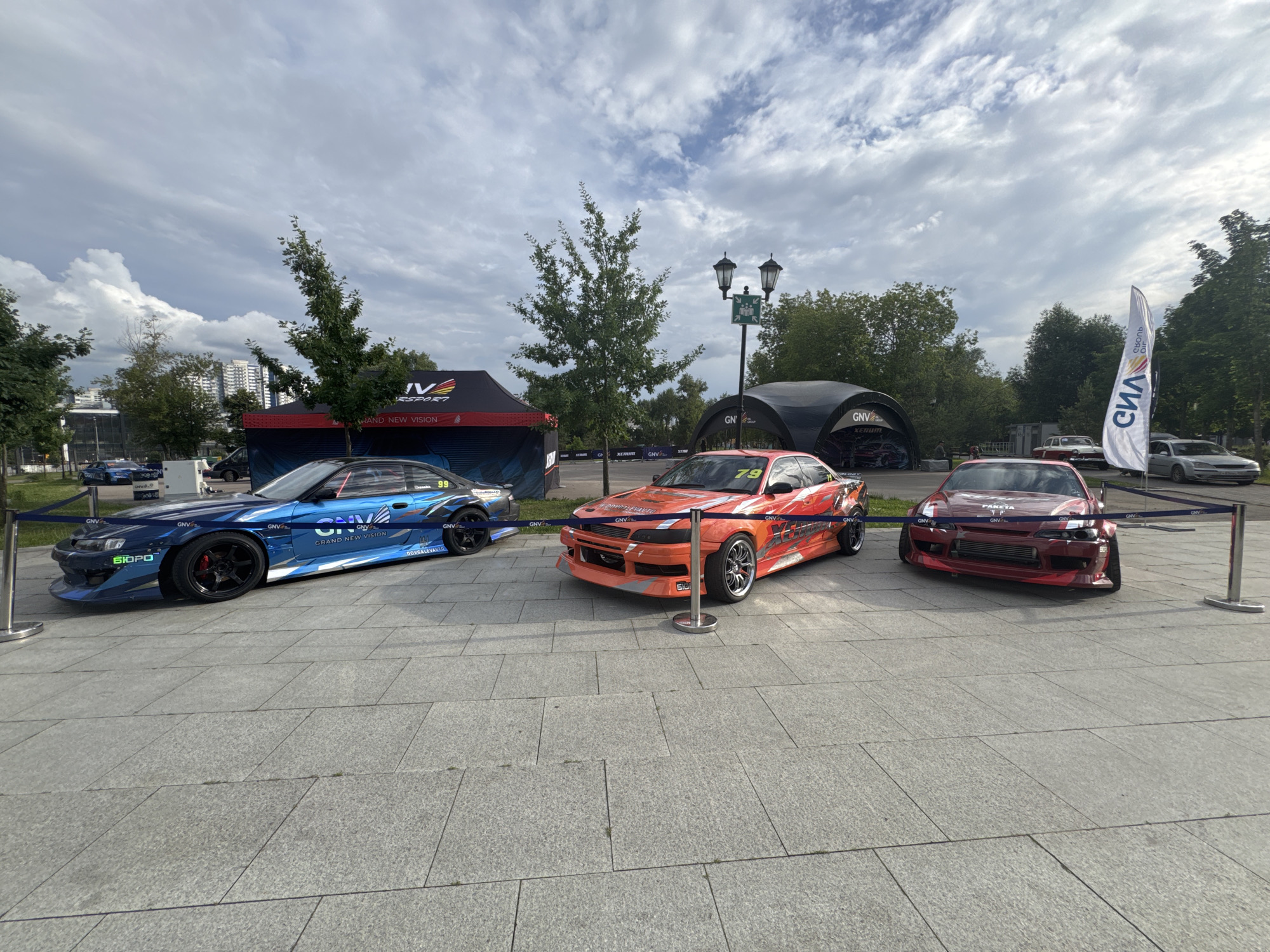 The image depicts an outdoor event with three modified sports cars parked in a row on a paved area. The cars are a blue Nissan Silvia S15, an orange Nissan Silvia S15, and a red Nissan Silvia S15. The blue car is on the left, the orange car is in the middle, and the red car is on the right. All three cars are heavily customized with aerodynamic body kits and racing decals.
Behind the cars, there are tents and structures associated with a motorsport event. One tent is red and black with "IV M...RSPORT" and "BRAND NEW VISION" visible on it. Another structure is a dark, curved tent with "GNV" logo. A white flag with Cyrillic text and a logo is also visible on the right side of the image. The background includes trees, buildings, and a cloudy sky.
The scene suggests a car show or a racing event, possibly a drift competition, given the nature of the modified cars. The paved area appears to be a public space, possibly a promenade or square in a city. The overall atmosphere is that of an organized outdoor event. Based on the visual cues, such as the architecture and the type of event, the location is likely in Moscow, Russia.