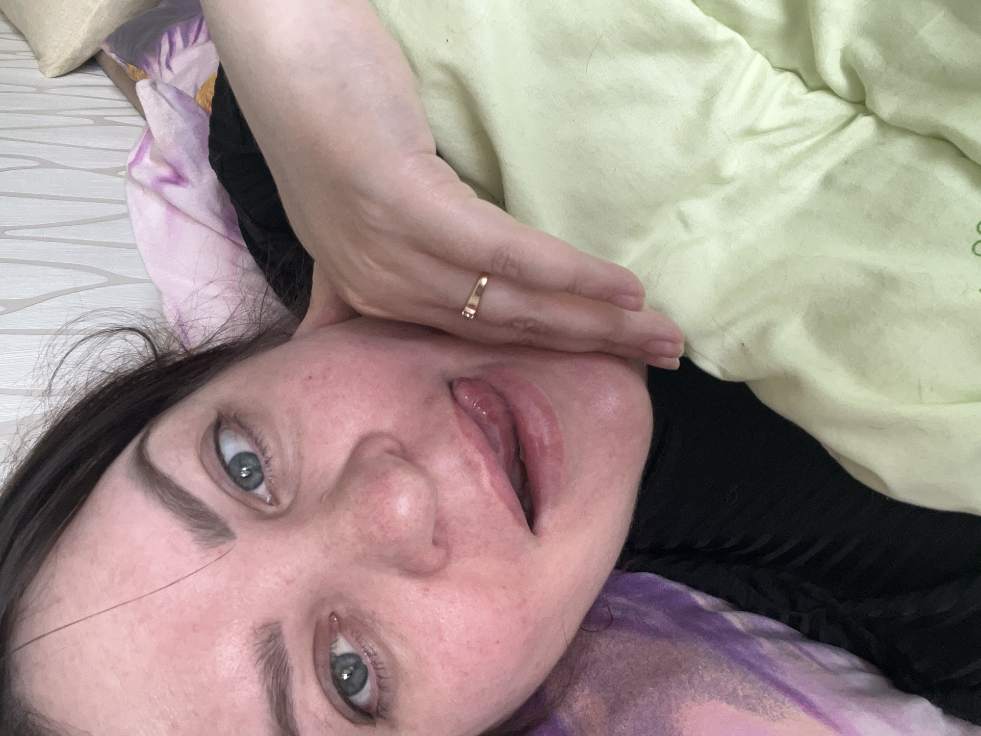The image presents a close-up, low-angle selfie of a woman lying down, looking directly at the camera. Her head is tilted, and her long, dark hair is visible towards the bottom left. She has striking light blue-grey eyes and dark, well-defined eyebrows. Her mouth is slightly agape, revealing the inside of her upper lip. Her left hand, adorned with a simple gold-colored band ring on her ring finger, is positioned near her jawline.
The woman appears to be resting on or under various fabrics. A light green blanket or garment covers a significant portion of her upper body, displaying some wrinkles and a faintly visible, illegible green text or logo in one section. Below her, a purple patterned sheet or fabric is visible, along with a dark, possibly black, garment. A plain, light-colored pillow is partially seen in the upper left corner, suggesting an indoor setting like a bedroom. The lighting is soft and ambient, providing no specific clues about the time of day or external weather conditions. The overall mood conveyed is one of relaxation or a casual, intimate moment. This scene is set in Perm, Russia.