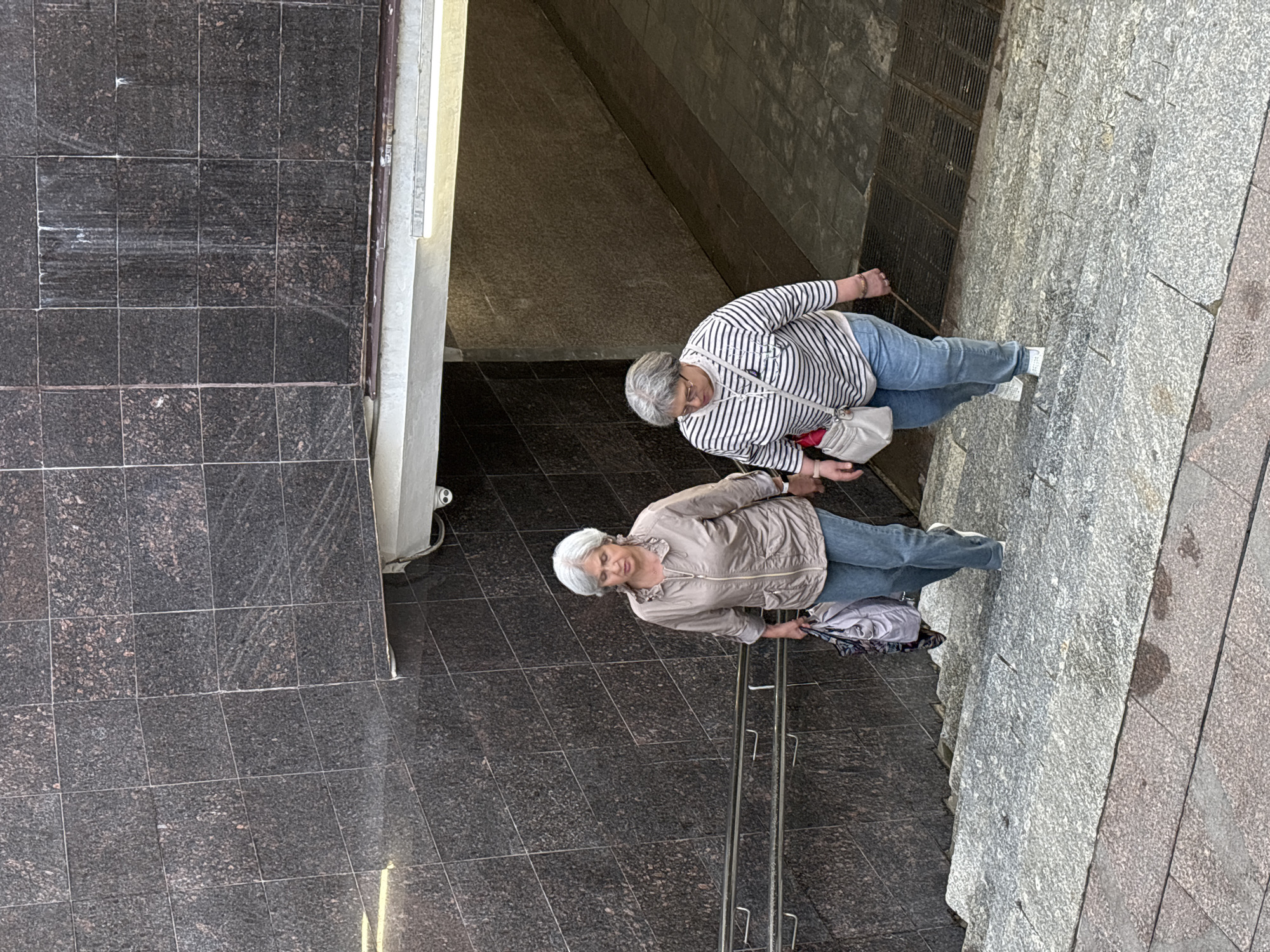 Two elderly women are walking down a stone staircase in what appears to be an underpass or subway station. The woman on the left has short gray hair and is wearing a light brown zipped jacket and blue jeans. She is carrying a patterned bag in her left hand. The woman on the right has gray hair and is wearing a striped long-sleeved shirt and blue jeans. She has a light-colored crossbody bag. The staircase is made of rough-hewn granite, and the walls are tiled with dark granite. A handrail is visible on the left side of the stairs. Above the staircase, a bright light illuminates the entrance to a tunnel. A security camera is mounted on the ceiling of the tunnel entrance. The overall environment is somewhat worn and utilitarian, consistent with public infrastructure. The context of Nizhny Novgorod, Russia, suggests this could be a typical urban underpass.