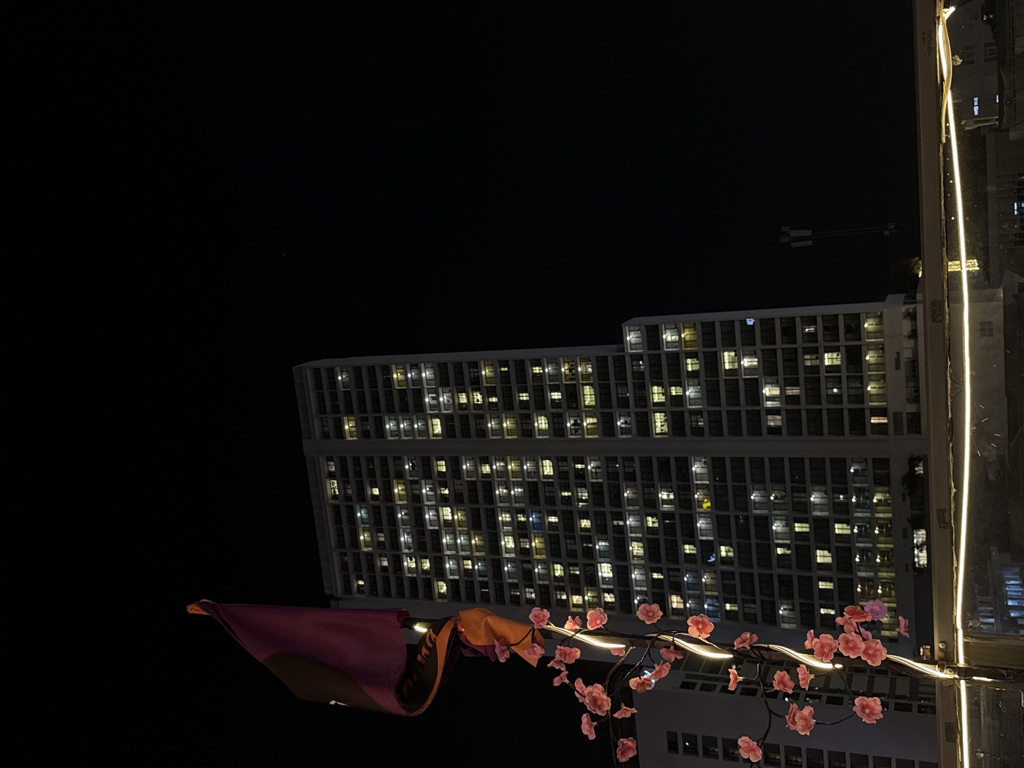 The image shows a night scene in Nha Trang Ward, Vietnam, focusing on a building facade with illuminated windows against a dark sky. A decorative display is in the foreground, featuring artificial pink flowers strung along a light, with a maroon and orange draped cloth adjacent. The building appears to be a high-rise, constructed in a grid pattern of windows. A crane is visible in the distance. The light from the display illuminates the scene. The image is taken from an elevated perspective.