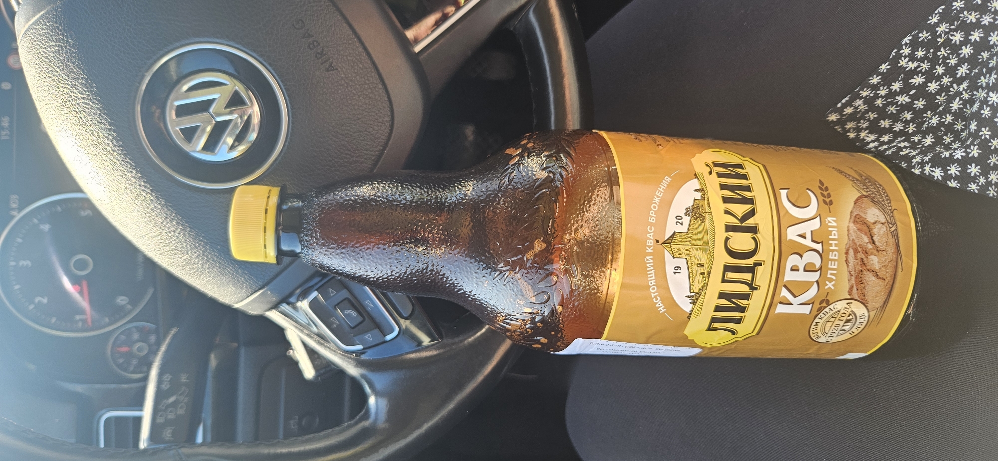 A brown glass bottle of "Lidskiy Kvas Khlebny" sits in the driver's seat of a car. The car's dashboard and steering wheel are visible in the background, along with the car's infotainment system displaying "15:46". The bottle is topped with a yellow plastic cap. The label on the bottle features a drawing of a castle and wheat stalks, and text in Russian that translates to "Lidskiy Kvas Khlebny" (Lidskiy Bread Kvass). The car appears to be parked, possibly in a city or urban area, as suggested by the presence of car interior elements like a steering wheel and dashboard. The presence of a car interior suggests the photograph was taken inside the vehicle, likely in Poland, given the request to locate it in Terespol, Poland. However, the image itself provides no direct visual clues to confirm the exact location within Terespol or any specific city.