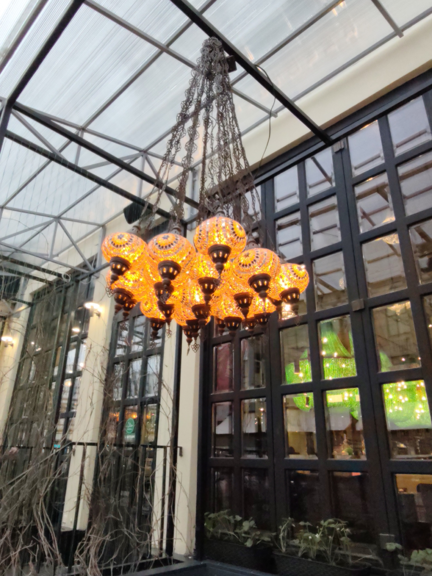 The image depicts an indoor space in Moscow, Russia. A decorative chandelier with orange mosaic glass globes hangs from the ceiling. The chandelier is suspended by chains. The ceiling is a grid of dark metal beams with transparent panels. Large windows with dark frames are visible on the right side of the image, reflecting interior lighting and greenery. On the left side, there are windows with bare branches in front of them. Potted plants are present below the windows. There are no people visible at the front of the shot, just the side of the head of a person on the left side.