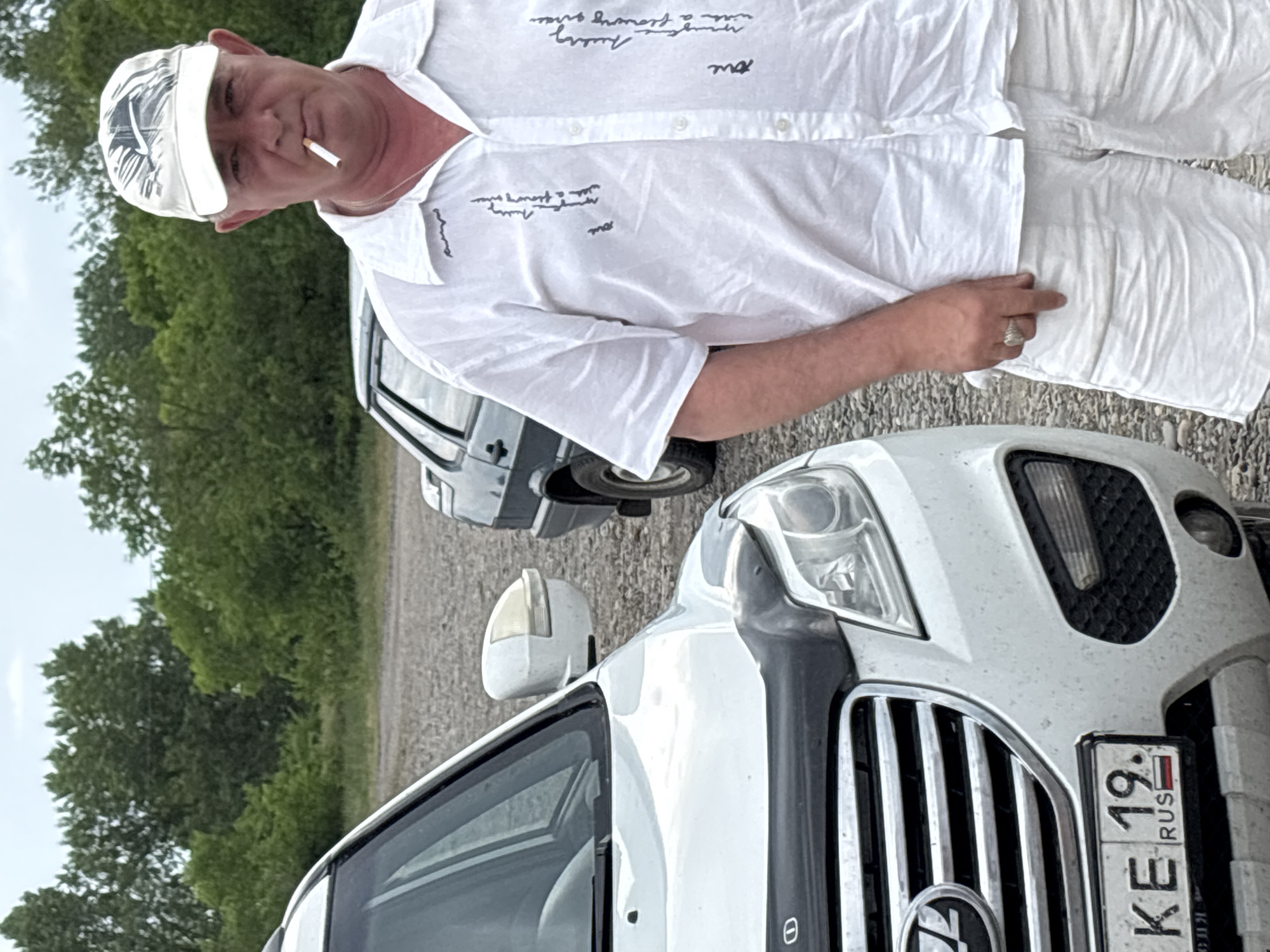 A man in a white shirt and shorts, wearing a white cap and smoking a cigarette, stands beside a white SUV. The man is looking at the camera. The SUV has a license plate that reads "KE 19. RUS". In the background, there are trees and another car, which appears to be a dark-colored sedan. The ground is covered with gravel. The overall setting suggests an outdoor, possibly rural or suburban location, in the city of Chernogorsk, Russia.