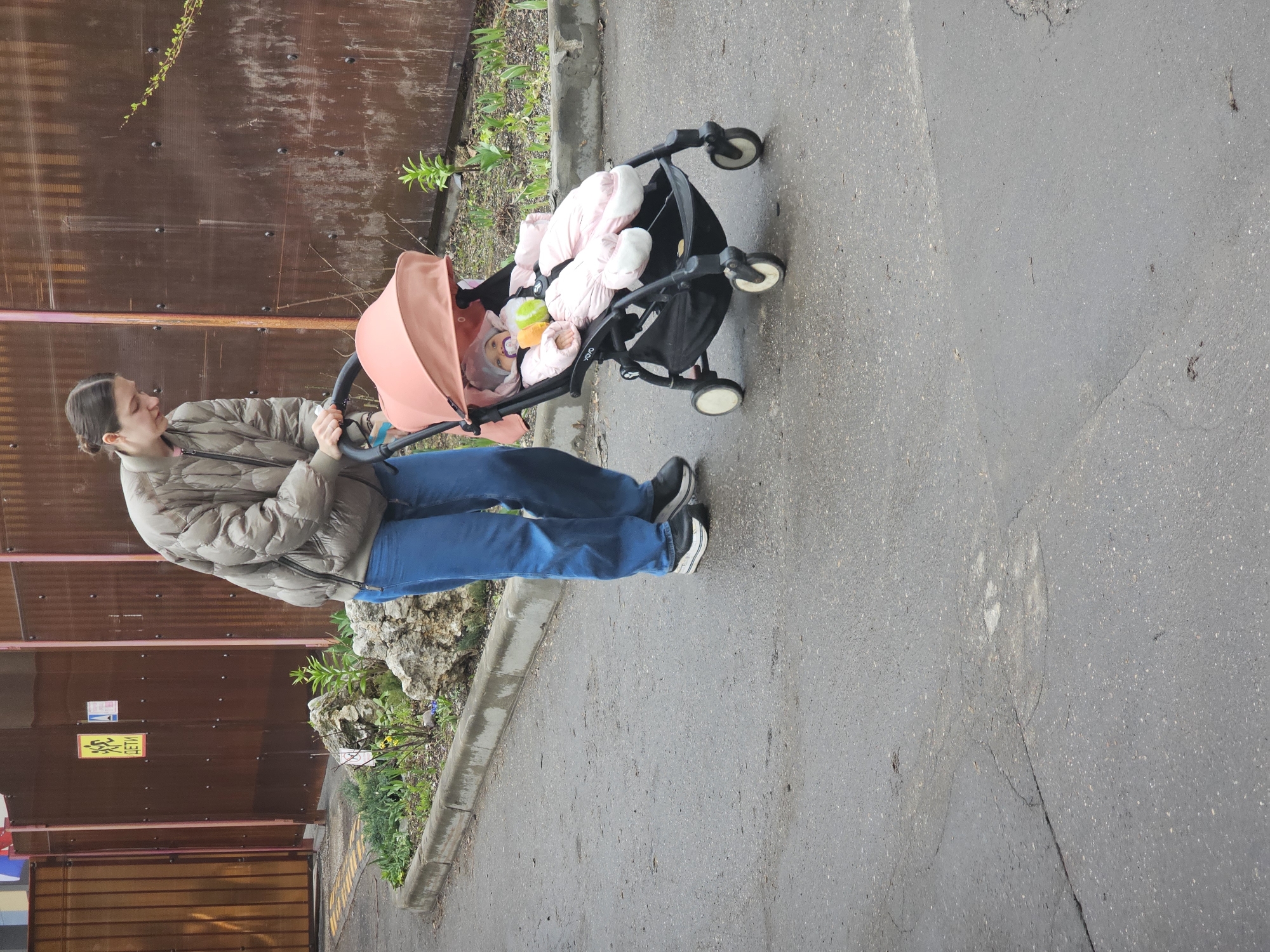 The image shows a woman pushing a baby stroller on a paved surface, likely a street or walkway. The woman is wearing a light-colored puffer jacket and blue jeans, with dark sneakers. The baby in the stroller is dressed in pink and has a peach-colored shade over the stroller. A toy is also visible in the stroller. The background includes a rusty brown corrugated metal fence. A curb with vegetation is also visible. The scene appears to be located in Moscow, Russia, as indicated by a yellow sign in the lower left with Cyrillic characters.