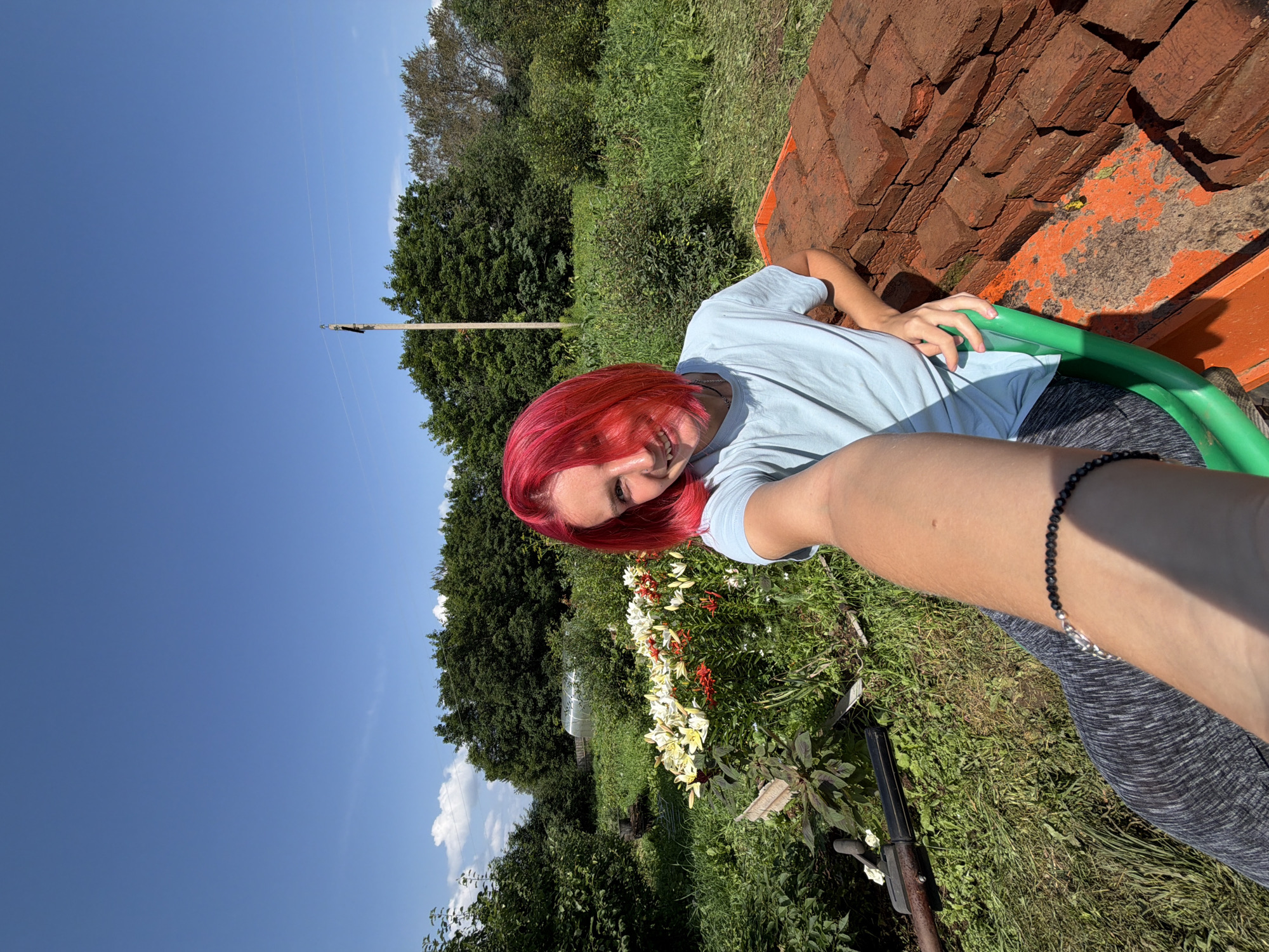 A young person with bright red hair takes a selfie in a sunlit garden. They are wearing a light blue t-shirt and dark grey leggings. Their left arm is extended, holding the camera. In the background, there are lush green trees, a greenhouse, and a patch of colorful lilies. To the right of the person, a pile of red bricks is visible. The sky above is a clear, bright blue. The location is the village of Zabolotovo, Russia.