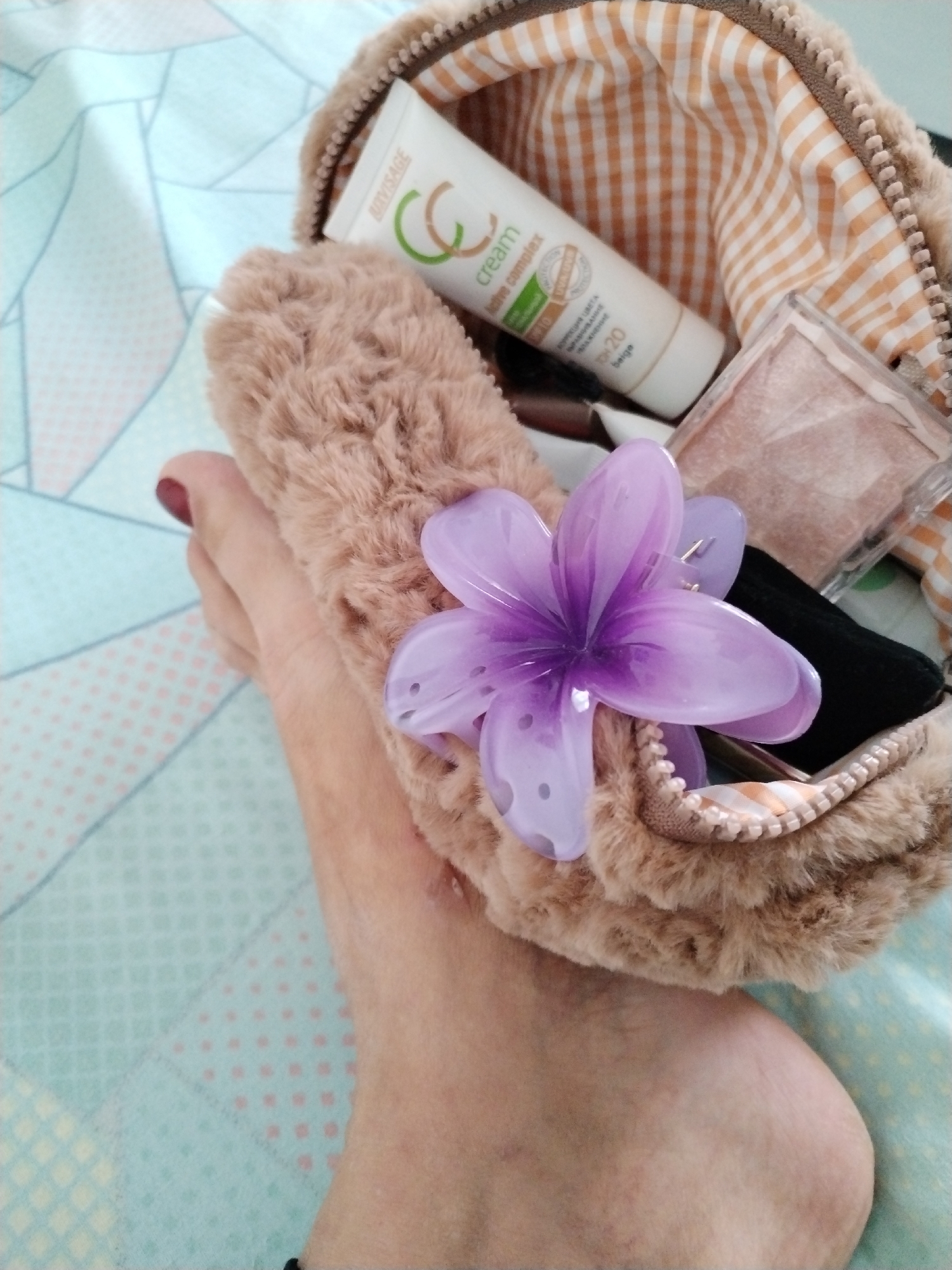 A close-up, overhead shot shows a person's foot holding a fluffy, light brown makeup bag. A translucent purple flower hair clip is attached to the top of the bag. Inside the bag, a tube of "Beliy Svet CC Cream" is visible, along with a compact of blush or powder. The background consists of a pastel blue and pink patterned blanket. The scene is likely indoors.
