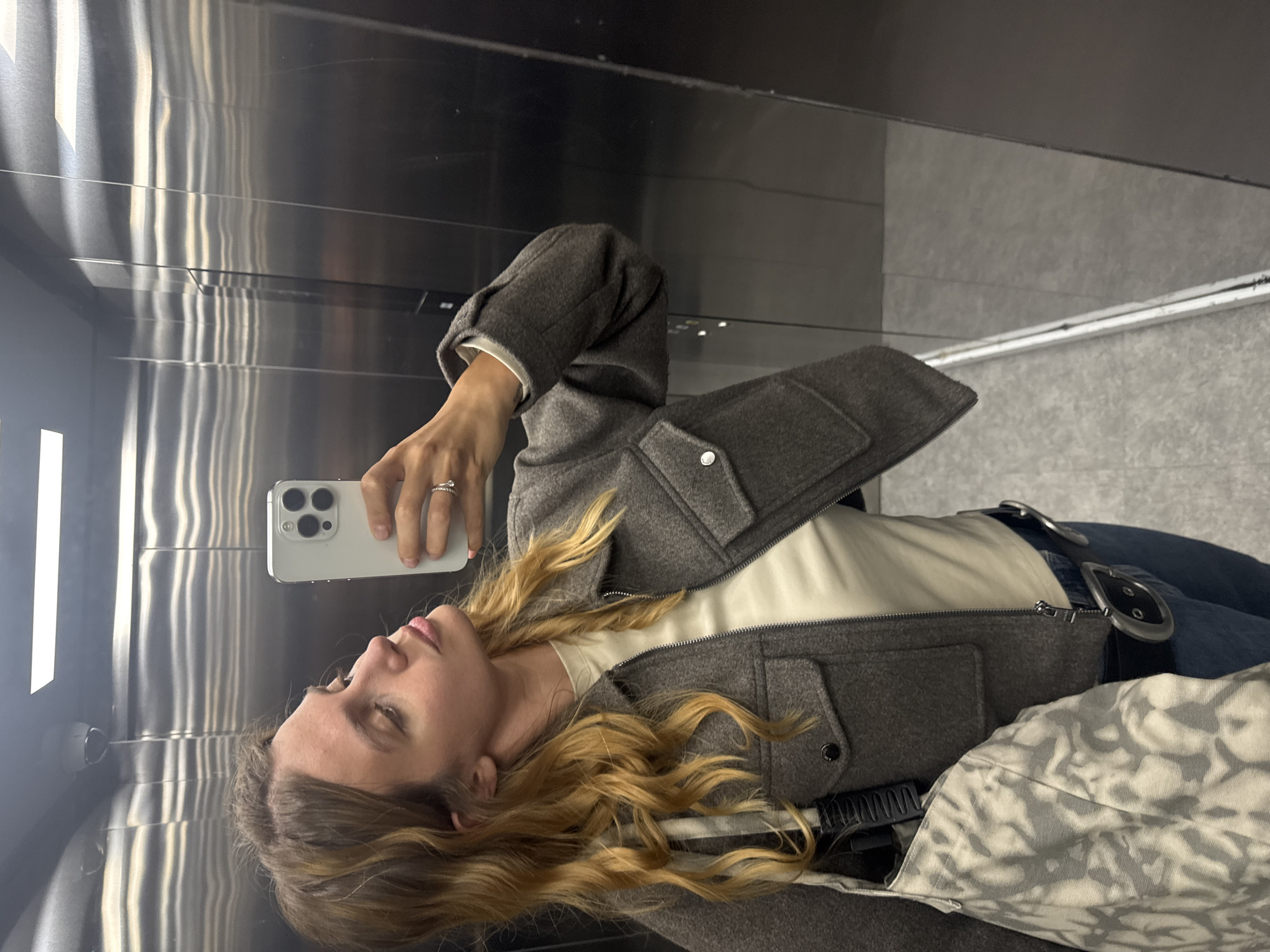 A person is captured in a selfie taken in a highly reflective surface, likely a polished metal wall or mirror, within what appears to be an elevator. The subject, a woman with long, wavy blonde or light brown hair, occupies the left side of the frame, looking slightly upwards and away from the camera.
She is dressed in a dark gray zippered jacket with two flap pockets secured by snap buttons, worn over a light cream or off-white top. She also wears blue jeans cinched with a black belt featuring a large, prominent silver buckle. Her right hand, adorned with a ring on the ring finger, holds a light-colored smartphone (possibly an iPhone with a triple-lens camera array) to capture the image.
The background is dominated by the reflective, possibly brushed metallic, walls of the enclosed space, displaying vertical streaks of light from unseen sources. A horizontal light fixture is also visible in the upper part of the reflection. At the bottom right, a portion of a patterned bag or fabric with an abstract grey and white design is visible. The scene is indoors, offering no cues about the time of day or weather. No text is visible in the image.
