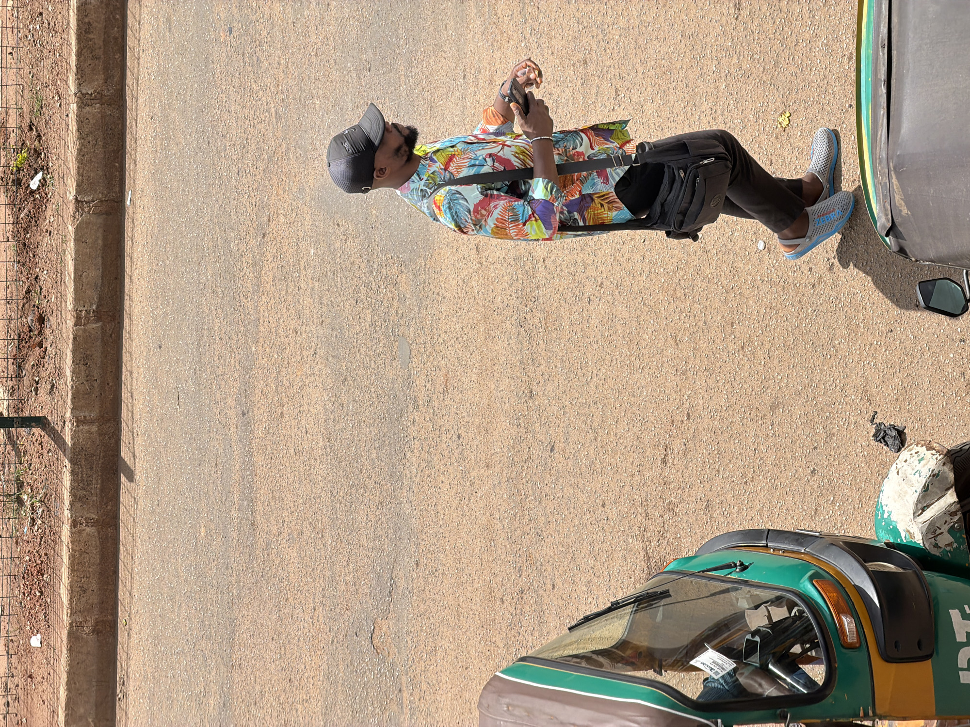 A man wearing a black baseball cap and a colorful, tropical-print shirt is standing on a dusty road in Bukuru, Nigeria. He has a beard and is looking down at a smartphone in his hands, engrossed in its screen. He is wearing dark pants and light gray clogs. A black satchel is slung across his chest. The scene appears to be outdoors during the day, with warm lighting casting shadows. To the right of the man, a portion of a green and yellow auto-rickshaw is visible, including its windshield and a side mirror. The road is paved with asphalt and shows signs of wear. The background on the left shows a dirt embankment with some sparse vegetation, bordered by a metal fence and a concrete structure. The overall impression is of a casual, everyday moment captured in a Nigerian street setting.