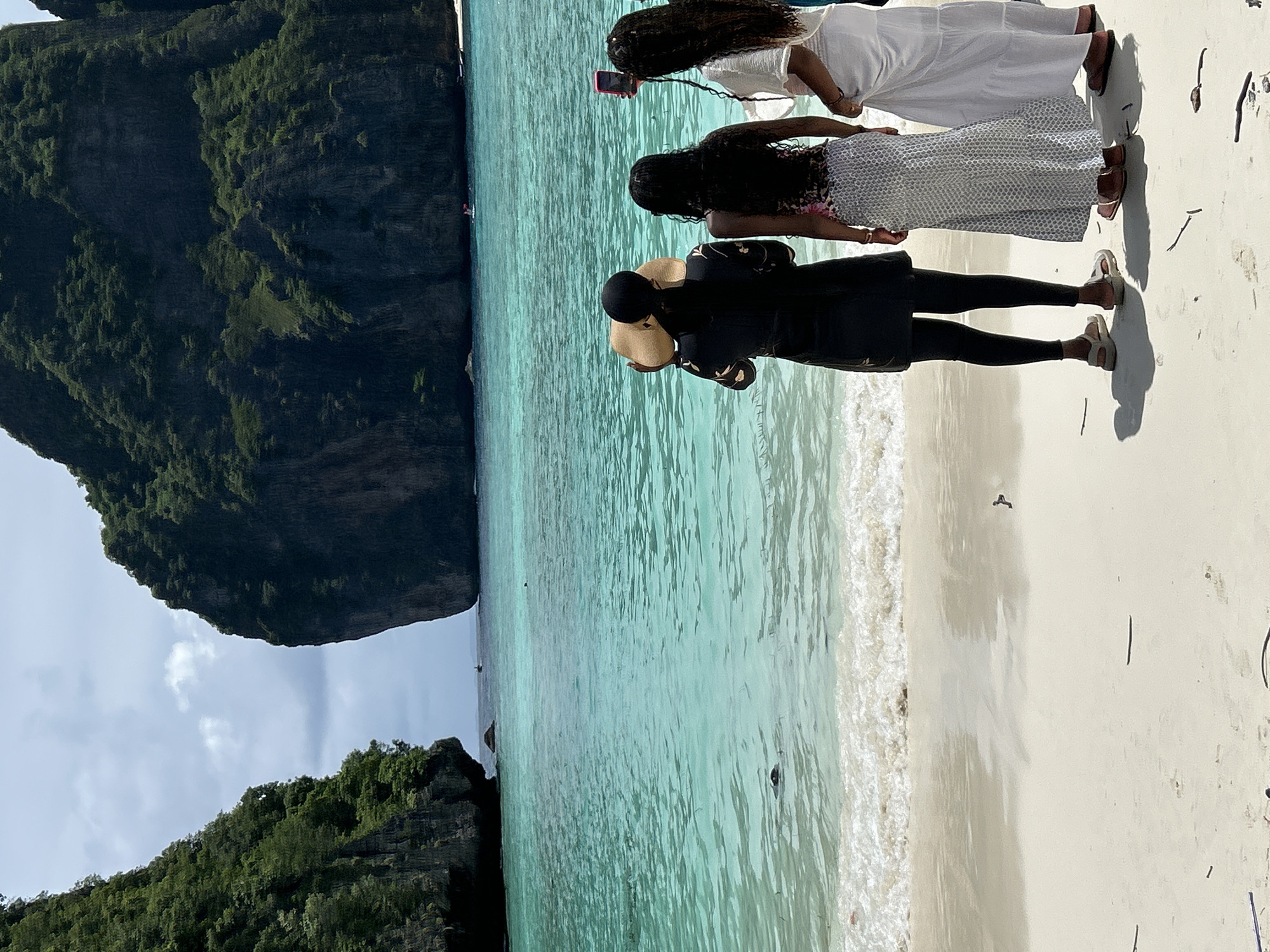 The image depicts Maya Bay in Ao Nang, Thailand, a picturesque location renowned for its stunning limestone cliffs and turquoise waters. In the foreground, three individuals stand on a white sandy beach, facing the ocean. The person on the left is wearing a black dress and a wide-brimmed hat, with their back to the camera. The middle individual has long dark hair and is wearing a patterned top and a long skirt. The person on the right, also with long dark hair, is wearing a white top and skirt and is holding up a pink smartphone, possibly taking a photo. The water is a vibrant turquoise, with gentle waves lapping at the shore. In the background, the iconic towering limestone cliffs of Maya Bay dominate the scene, their green foliage contrasting with the blue sky. Several small boats are visible in the distance, and a lone swimmer is near the center of the bay. The overall atmosphere is serene and idyllic, characteristic of a tropical paradise.
